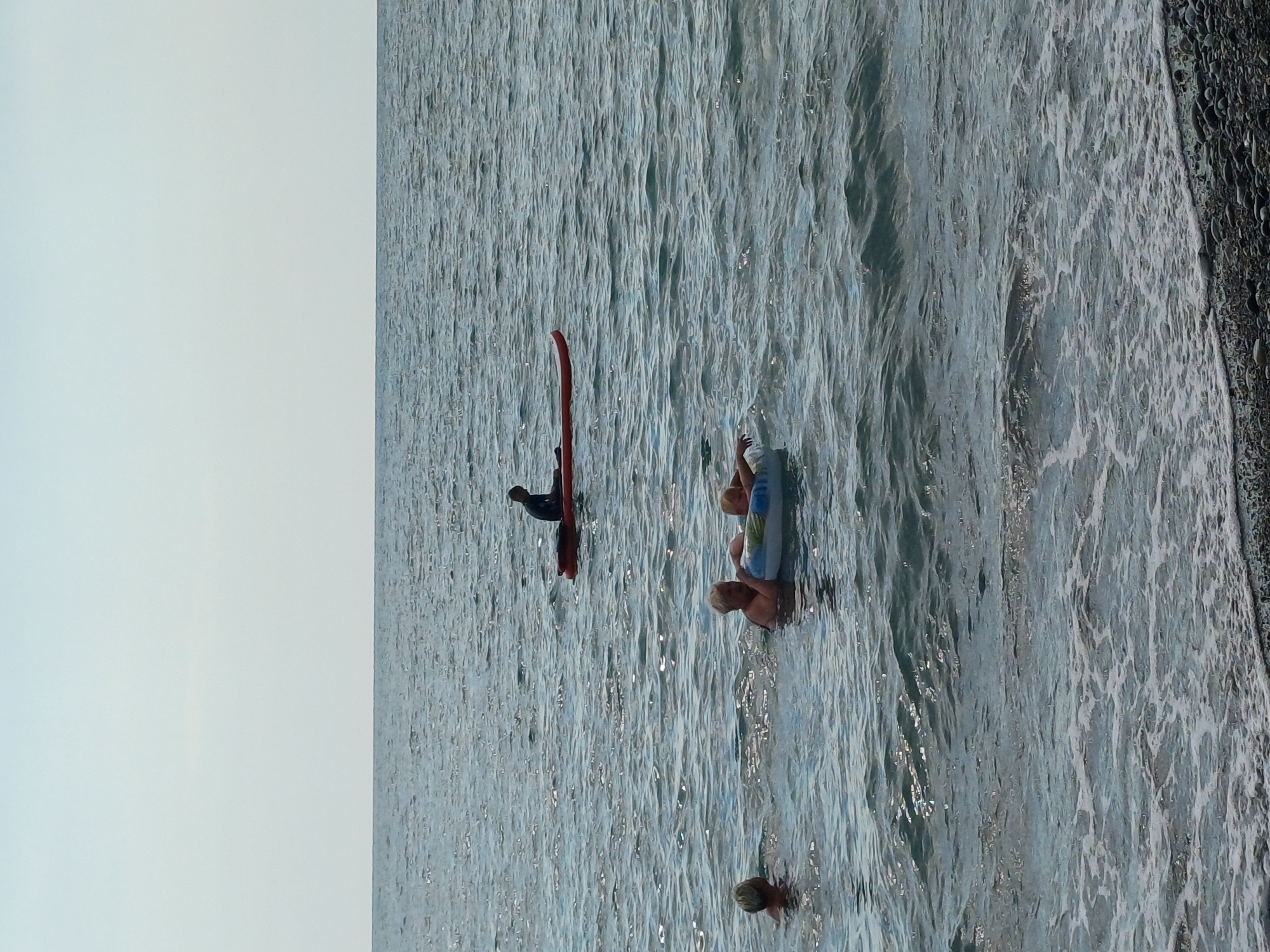 In this vertically oriented image, several individuals are present in the ocean near a rocky shore. The scene appears to be a beach or coastal area, with the water exhibiting a textured, wavy surface that catches the light, suggesting it might be midday under sunny or bright conditions.
The subjects include at least three people. One person is lying on a blue and white inflatable raft, appearing to relax in the water. Another individual is in the water, possibly swimming or wading, with only their head visible. A third person is seen in the distance, next to a bright red object that resembles a surfboard or paddleboard. This individual is dark-haired and is positioned as if interacting with the red object. There are also hints of other people further out in the water, suggesting a recreational activity is underway.
The water is a light blue-grey, with visible ripples and small waves. The rocky shore to the right of the image is dark and wet, with distinct textures indicating pebbles or stones. The sky is a pale, bright blue, taking up a significant portion of the left side of the image. No text is visible in the frame. The overall atmosphere seems relaxed and casual, indicative of people enjoying a day at the beach. Given the location context of Sochi, Russia, this could be a scene from the Black Sea coast.