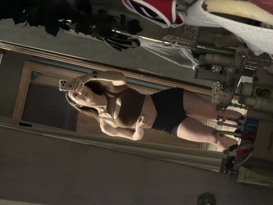 A woman takes a mirror selfie. She wears a brown, strappy sports bra and black shorts. The mirror shows a cluttered space, including a storage rack with containers and items on it. There's a window frame visible in the reflection, as well as objects hanging from the ceiling above, including what appears to be a helmet or hat. The room has a slightly dark and cluttered appearance.