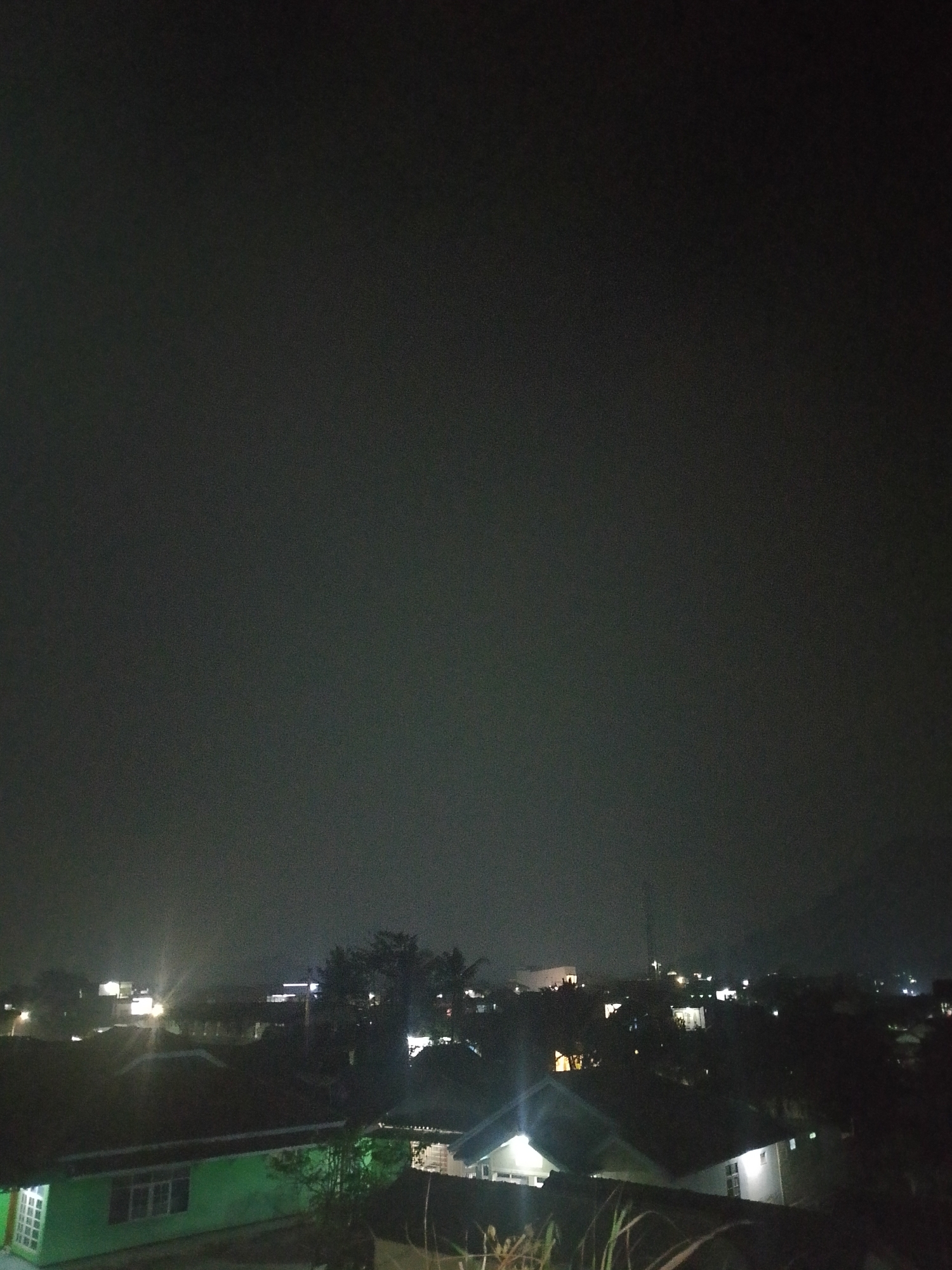 The image depicts a nighttime aerial view of a residential area in the city of Situwangi, Indonesia. The sky is dark, indicating nighttime, with a slight haze or cloud cover. The scene is illuminated by artificial lights from houses and streetlights. Several houses with visible rooftops are scattered throughout the frame. Some houses have lit windows, suggesting occupancy. Trees and vegetation are present, appearing as dark silhouettes against the illuminated background. In the distance, more lights indicate the presence of a larger settlement or city. The overall impression is that of a quiet, suburban or semi-urban environment at night.