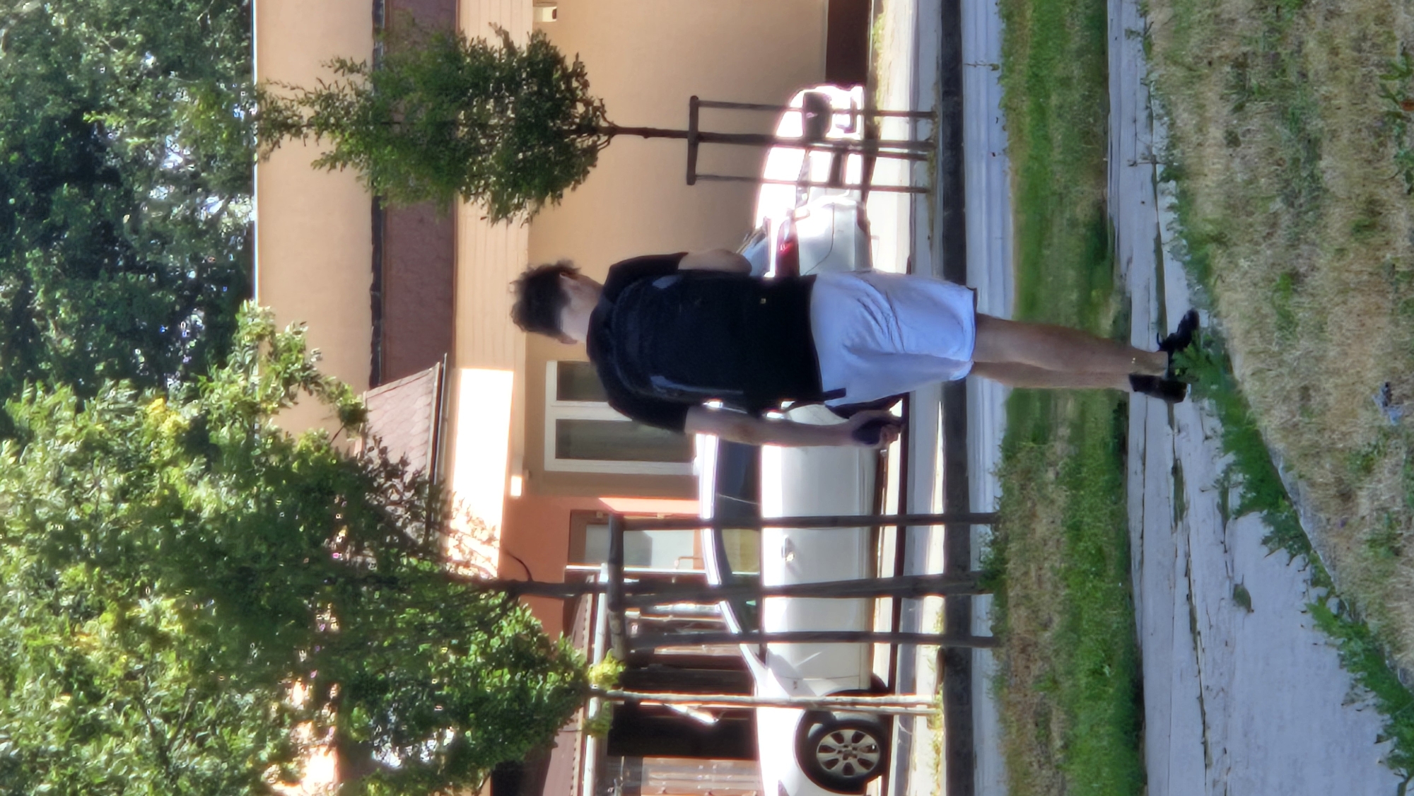 In Prešov, Slovakia, a young man with dark, curly hair walks away from the camera on a paved path. He is wearing a black t-shirt and white shorts, with a black backpack on his back. He holds a dark object, possibly a phone, in his right hand. The path is bordered by a strip of dry grass on the right and a patch of greener grass on the left. In the background, a white car is parked in front of a modern building with light-colored walls and a dark roof. Two small, potted trees stand on either side of the path, and larger trees with green foliage are visible behind the building. The overall scene appears to be a bright, sunny day, suggesting it is likely springtime or summer.