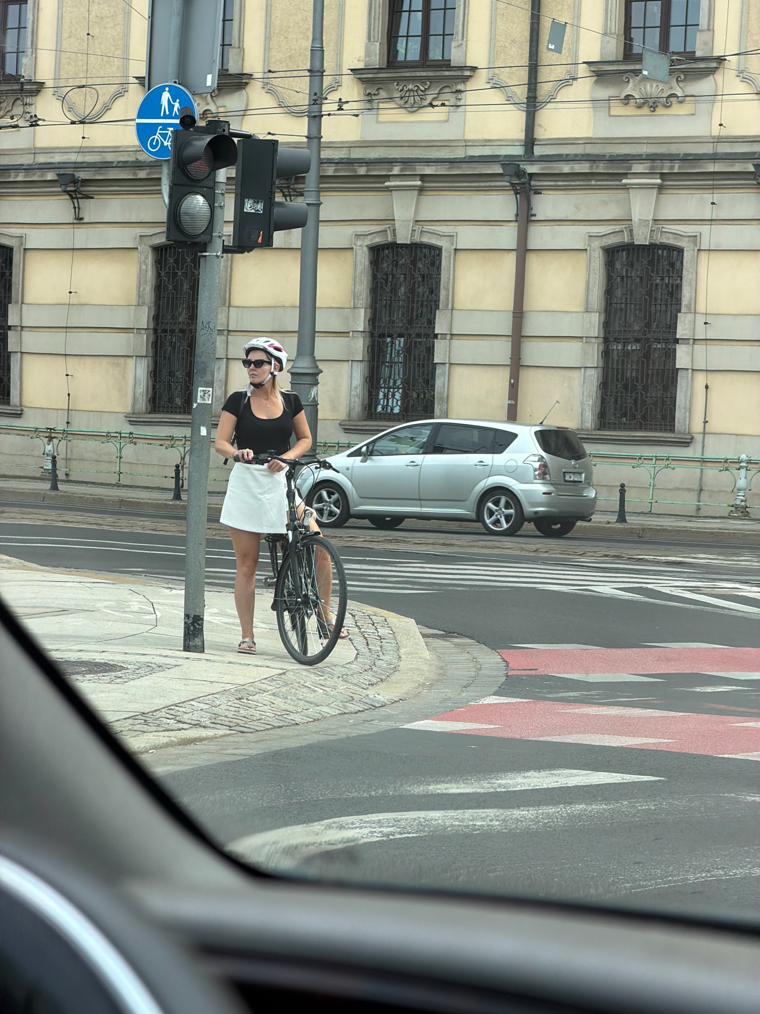 A woman wearing a white helmet, sunglasses, a black t-shirt, and a white skirt stands with her bicycle on a sidewalk next to a traffic light pole in Wrocław, Poland. The traffic light pole has a blue sign with a pedestrian and bicycle symbol. Behind her, a silver van is parked on the street, and a building with arched windows and decorative elements is visible. The sidewalk is paved with cobblestones, and a crosswalk with red and white stripes is on the street. The scene is viewed from inside a car.