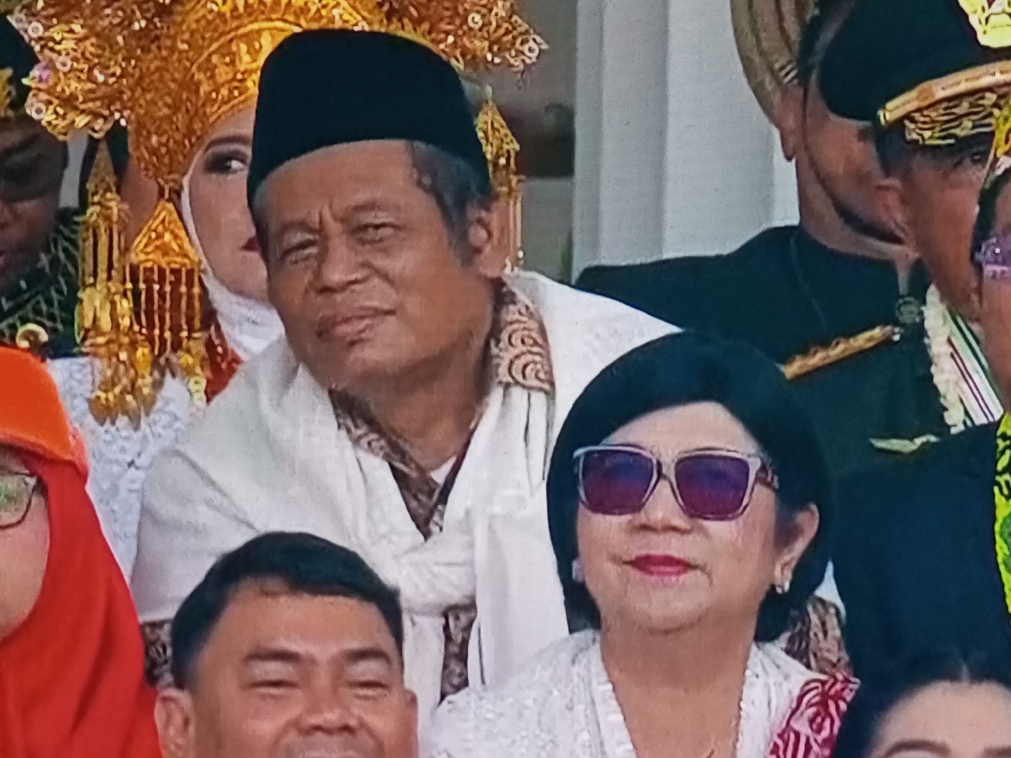 In the city of Banyudono, Indonesia, a group of individuals are gathered, likely for a cultural event or ceremony, as indicated by the traditional attire. A man in the foreground, wearing a black peci cap and a white garment with ornate patterns, is positioned centrally. To his left, a woman with a very elaborate golden headdress, possibly a bride, is partially visible. To the right of the man, a woman wearing sunglasses with purple lenses and a white lace top is also present. In the lower portion of the image, a man with short hair is looking towards the viewer with a wide smile. The background appears to be an indoor or covered outdoor setting, with a plain white wall or surface dominating the upper right portion of the image. The overall impression is one of a formal gathering with a focus on cultural heritage.