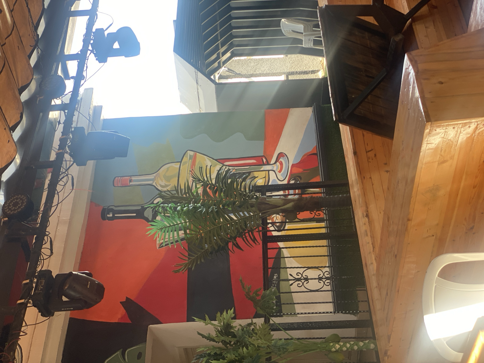This image depicts an outdoor or semi-outdoor commercial establishment located in Howayi, Nigeria, under bright daylight conditions. An overhead metal truss system supports multiple professional stage lighting fixtures, including a "BEAM" moving head light and an LED par light, with associated wiring visible against a corrugated, brownish ceiling. A large, colorful mural covers the left wall, featuring abstract geometric shapes in red, black, green, yellow, and light blue, with prominent outlines of two bottles and a martini glass with a green garnish. Two artificial palm trees stand in the mid-ground, positioned in front of a black wrought-iron railing with decorative scrollwork. The foreground consists of light-colored wooden plank structures, suggesting seating or bar elements. In the far right, a dark, modern building with a ribbed exterior is visible, partially obscured by strong sunlight, with stacked white plastic chairs and a tilted dark wooden table in its vicinity. No individuals are present in the scene.