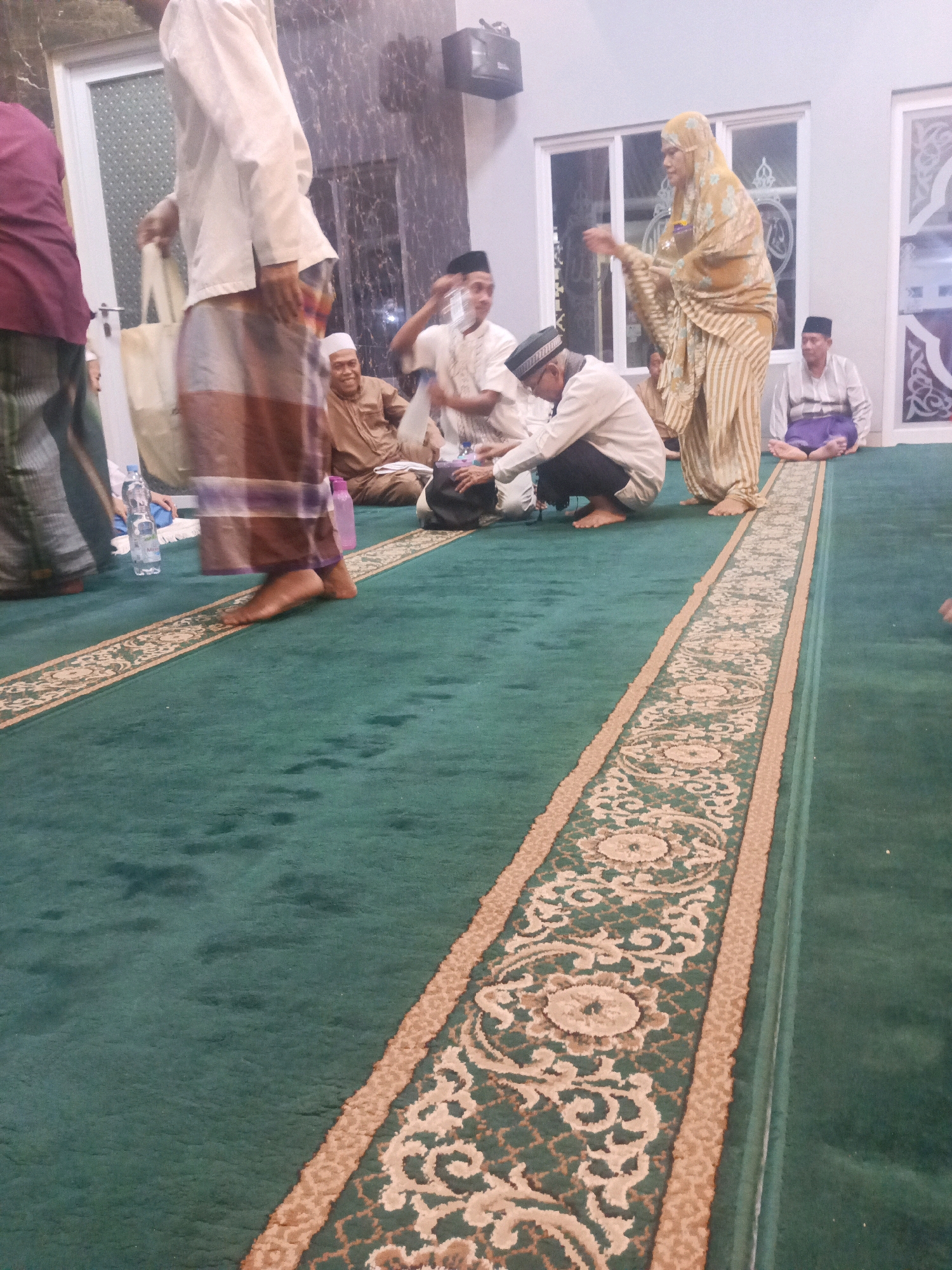 In a mosque in South Tangerang, Indonesia, a group of men and one woman are gathered on a green carpet adorned with a patterned runner. Some men are seated, while others are standing or in the process of sitting down. One man in the foreground, wearing a white shirt and patterned sarong, is carrying a tote bag. A woman in a yellow patterned hijab and dress is speaking to a seated man. Bottles of water are visible on the carpet. The environment appears to be indoors, with walls and windows suggesting a mosque interior.