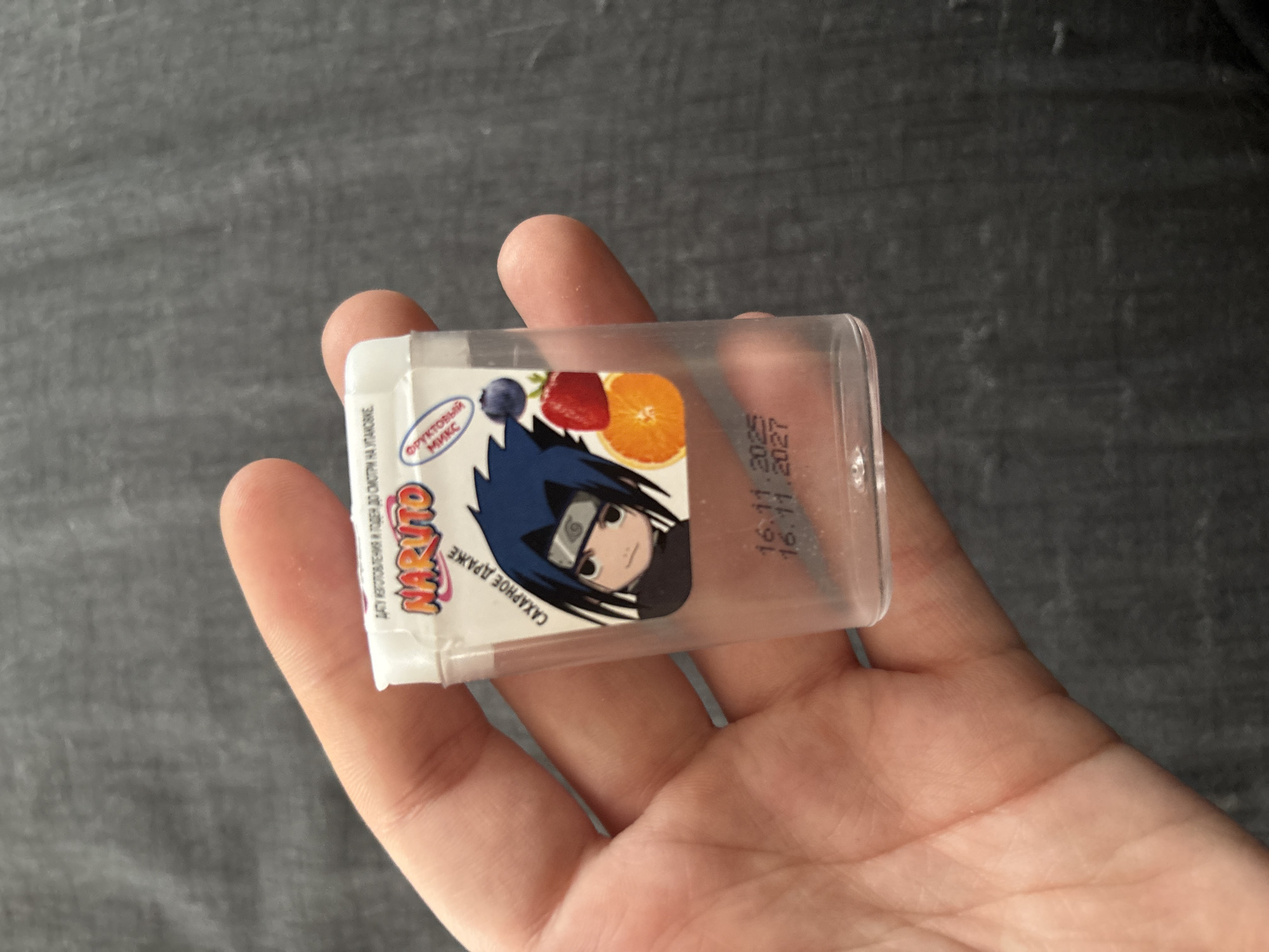 An indoor, close-up shot features a light-skinned hand holding a small, rectangular, transparent plastic container against a dark, mottled background. The container is oriented horizontally in the palm.
The container's white label displays the animated character Sasuke Uchiha from the "Naruto" series, along with illustrations of a blueberry, a strawberry, and an orange, indicating a "ФРУКТОВЫЙ МИКС" (Fruit Mix) flavor. The brand name "NARUTO" is prominently displayed. Other Russian text on the label includes "ДАТУ ИЗГОТОВЛЕНИЯ И ГОДЕН ДО СМОТРИ НА УПАКОВКЕ" (Date of manufacture and best before see on packaging) and "САХАРНОЕ ДРАЖЕ" (Sugar Dragee), suggesting the product is candy or dragees.
Printed directly onto the clear plastic of the container are two dates: "16.11.2025" and "16.11.2027". These likely represent the production and expiry dates, respectively. The background is a blurred, dark grey or black surface, possibly a tabletop, providing no further specific location cues beyond the general indoor setting in Khimki, Russia. The image focuses entirely on the hand and the candy container.