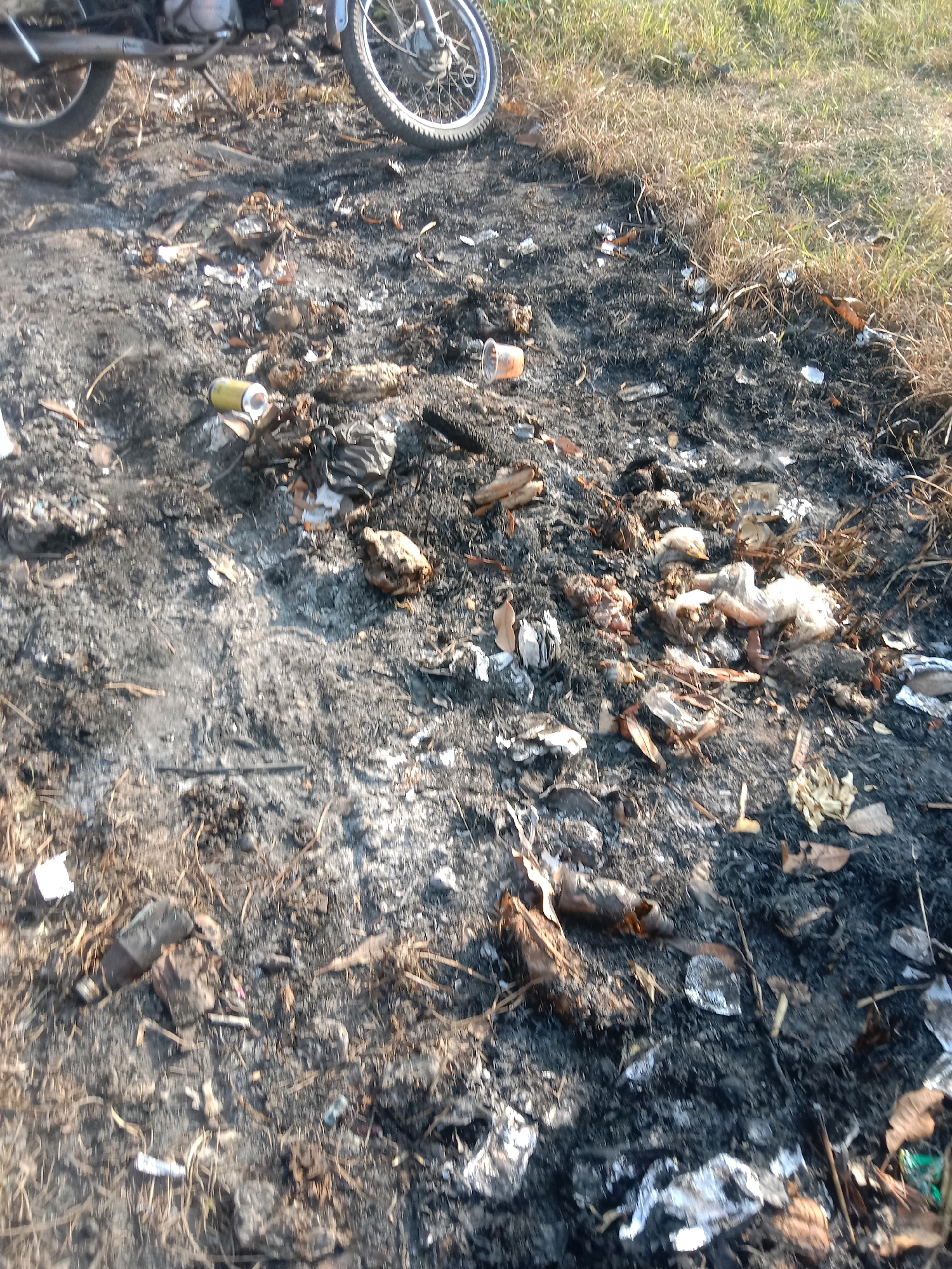 The image depicts a post-fire scene in Banjardowo, Indonesia. The ground is covered in ash, burnt debris, and scattered trash. Partially visible in the upper left corner is the wheel and exhaust pipe of a motorcycle, suggesting its presence at the scene. Amongst the ash and debris, remnants of plastic bottles, crumpled paper, and other unidentifiable burnt materials are visible. The surrounding environment is a grassy area, with dry, sun-bleached grass bordering the burnt patch. The lighting suggests it is daytime, possibly late afternoon due to the shadows. There are no people visible in the image. The scene appears to be an outdoor area, likely a roadside or an undeveloped plot of land.