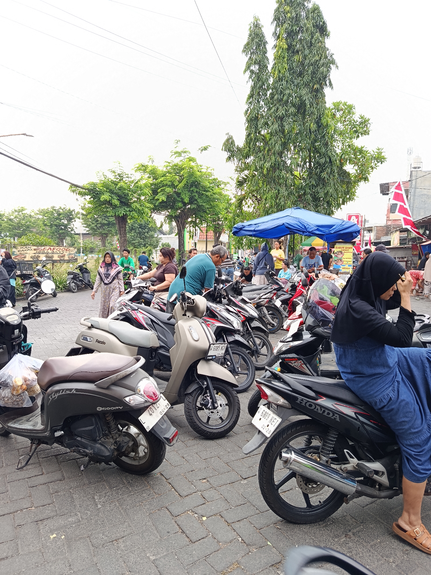 The image depicts a street scene in Keboansikep, Indonesia, likely a market area given the presence of stalls and a variety of parked motorcycles. In the foreground, several scooters and motorcycles are parked on a paved surface. One scooter, labeled "Fazzio" and "Scoopy," is prominently displayed, carrying a plastic bag filled with bread or pastries. Another motorcycle, a black Honda, has a person wearing a blue dress and a black hijab seated on it, adjusting their headscarf. Behind these vehicles, a blue tent is set up, under which several people are gathered. A woman in a patterned headscarf and dress walks through the scene, passing by parked motorcycles. More motorcycles and people are visible in the midground and background, with trees and buildings lining the street. A red and white flag is visible on a building in the distance. The overall atmosphere suggests a busy daytime commercial environment.