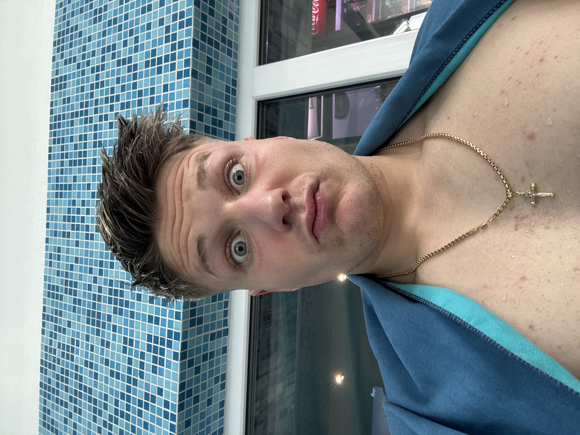 A close-up, tilted selfie shows a man with short, damp brown hair, light skin, and wide blue eyes. He is wearing a gold chain with a cross pendant and a blue hooded shirt or robe. His skin appears damp with noticeable pores and some blemishes.
Behind the man, a wall is covered in small, light blue mosaic tiles. To his right, a window or glass partition is visible. The reflection in the glass is distorted, but suggests an indoor setting.
While the image provides no direct indicators of location, the request specifically identifies it as Plovdiv, Bulgaria. Without additional context, confirmation of this location based solely on the visual content is not possible.