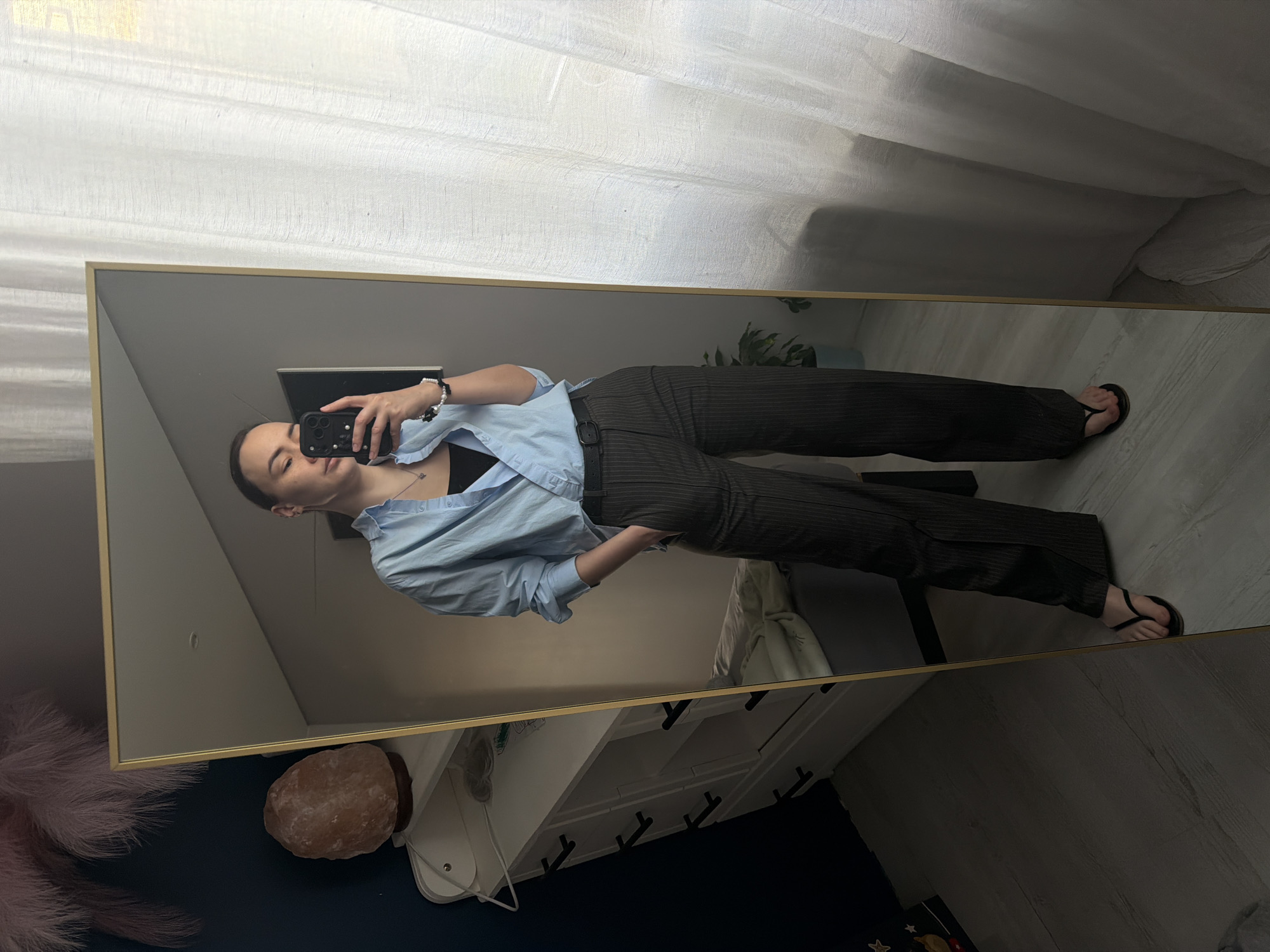 A woman is reflected in a mirror, taking a selfie. She is wearing a light blue button-down shirt over a black top, dark pinstripe pants, and black flip-flops. The mirror is framed in gold and reflects a room with white curtains, a white dresser with black drawer pulls, a salt lamp, and a pink feather duster. The location appears to be indoors, possibly a residential setting. There is no evidence to definitively place the location in Chaikovsky, Russia.
