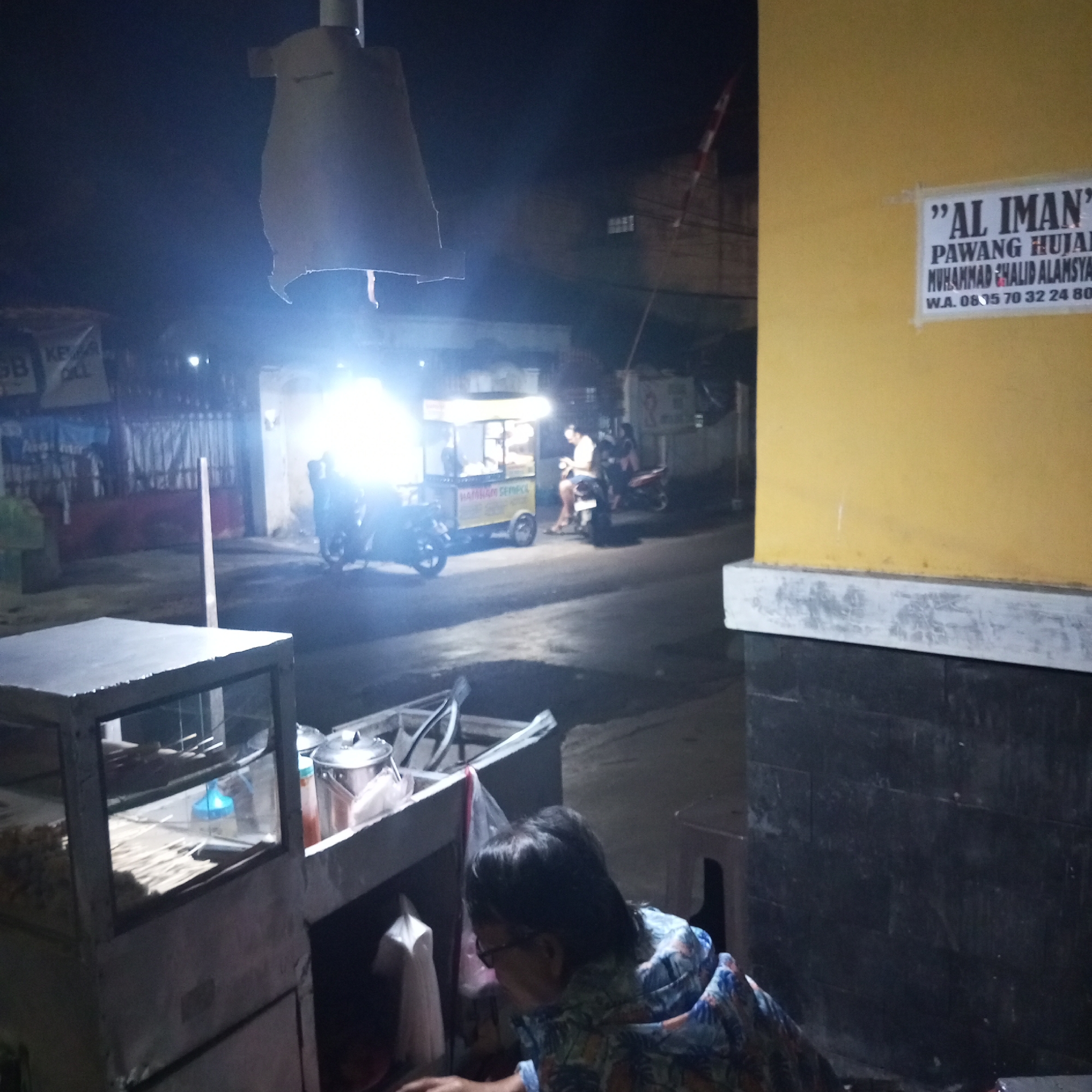 In the city of Sanggrahan, Indonesia, at night, a scene unfolds near a roadside food stall. A vendor, a woman with dark hair and glasses, is positioned behind a glass-fronted display case, which holds skewers of food and various containers. She appears to be attending to her work, with her body angled towards the display. In the background, a food cart illuminated by bright lights is visible, with a person sitting on a motorcycle next to it. Further down the street, other motorcycles and a food cart with a sign that reads "HAMHAM SEMPOL" are also present, indicating a busy area with food vendors. A yellow wall with a sign that reads "'AL IMAN' PAWANG HUJAN MUHAMMAD KHALID ALAMSYAH W.A. 0815 70 32 24 80" is prominent on the right side of the frame. The overall environment suggests a vibrant nighttime street scene in a commercial area.