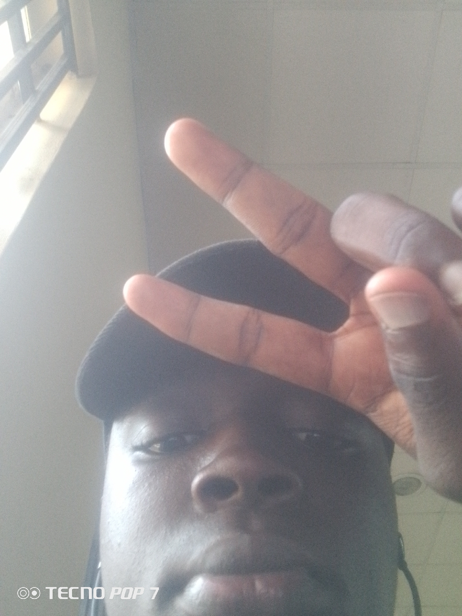 In Eleweran, Nigeria, a low-angle, close-up selfie is taken indoors, likely in a residential or public building. The subject, a person wearing a dark baseball cap, is positioned centrally, their face partially obscured by their own hand which is raised in a peace sign gesture. The background is a muted, neutral tone, possibly a wall, with a hint of a window or grate structure visible on the left side of the frame, indicating an interior space with natural light. The overall lighting is soft and diffused. The bottom left corner of the image displays the watermark "TECNO POP 7".