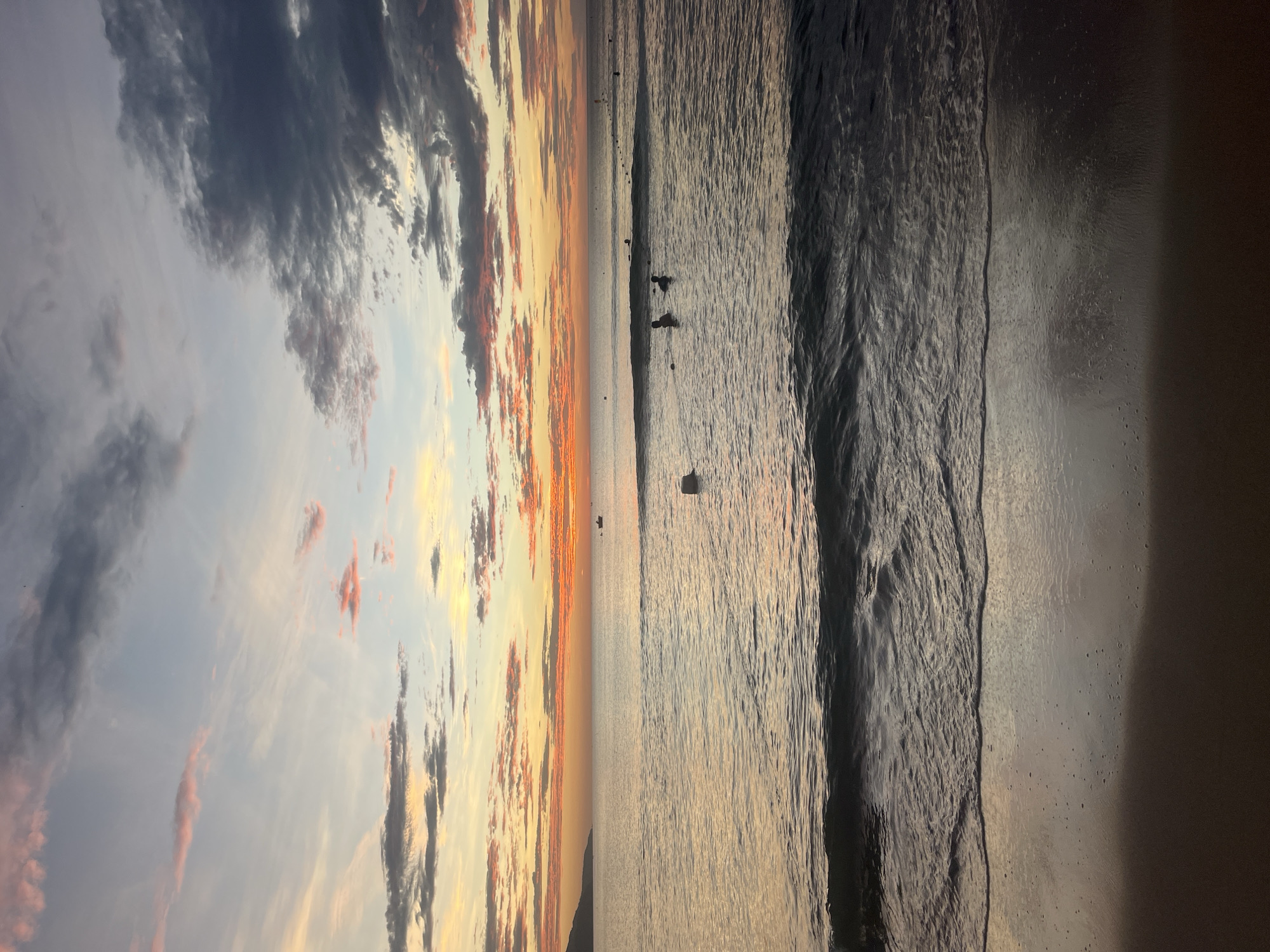 This media file presents a tranquil landscape captured at either sunrise or sunset, set in Đà Nẵng, Vietnam. The scene is dominated by a dramatic sky and a calm ocean, viewed from the sandy shore.
The upper portion of the image features a vast, cloud-filled sky. On the left, dark, heavy clouds create a somber tone, while towards the right, the sky is ablaze with warm hues of orange, yellow, and soft pink, illuminated by the low-lying sun. These vibrant colors streak across wispy and more substantial cloud formations.
A clear horizon line separates the expressive sky from the expansive ocean. The sea, occupying the middle ground, appears calm with gentle ripples across its surface, reflecting the muted light from above. Several small, dark silhouettes of people are visible in the water, suggesting they are wading or swimming. Two figures are more distinctly seen in the mid-distance, while other, much smaller figures dot the horizon line further out.
The foreground consists of the wet, dark sand of the beach, showing subtle textures from the ebbing waves and reflecting the ambient light. The overall atmosphere is one of serene natural beauty, with individuals enjoying the peaceful coastal environment during a picturesque dawn or dusk. No visible text is present in the image.