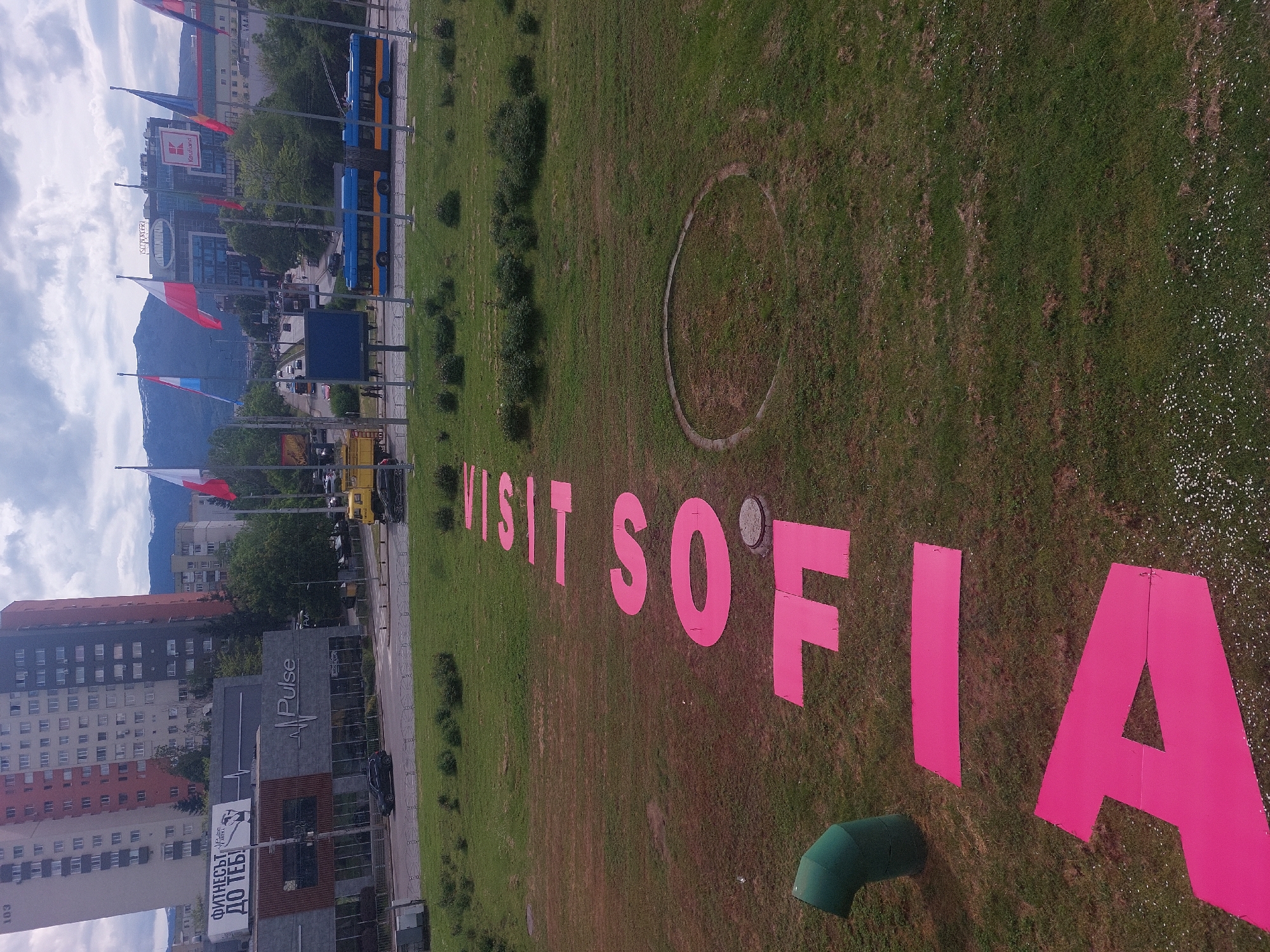The image depicts an aerial view of a grassy area where the words "VISIT SOFIA" are spelled out in large, bright pink letters. A circle is also visible in the grass. In the background, there is a street with buildings, flags, and a mountain range. The city is Sofia, Bulgaria, as indicated by the spelled-out word and the Cyrillic writing. The image is oriented in a non-traditional way and would require rotation for proper viewing. There are no people visible.