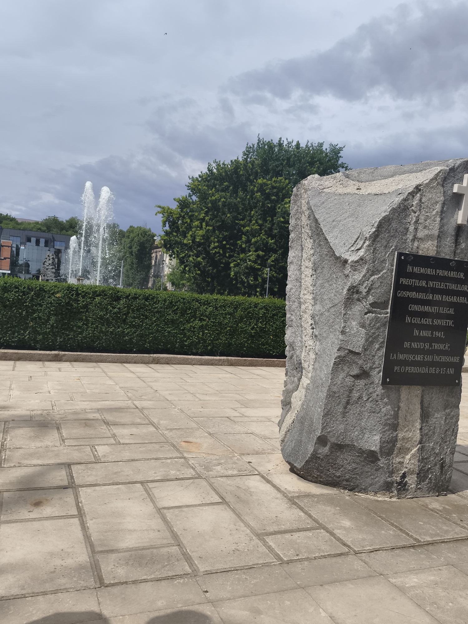 The image depicts a memorial monument in Chișinău, Moldova, located in front of a public fountain. The monument is a large, rough-hewn granite stone with a plaque attached. The plaque is inscribed with text in Romanian. To the left of the monument, a row of neatly trimmed green hedges lines the foreground. Behind the hedges, a multi-tiered fountain sprays water high into the air. In the background, modern buildings and lush green trees are visible. The ground in front of the monument and hedges is paved with large, square concrete tiles. The sky above is overcast with some dramatic clouds. There are no people visible in the image.