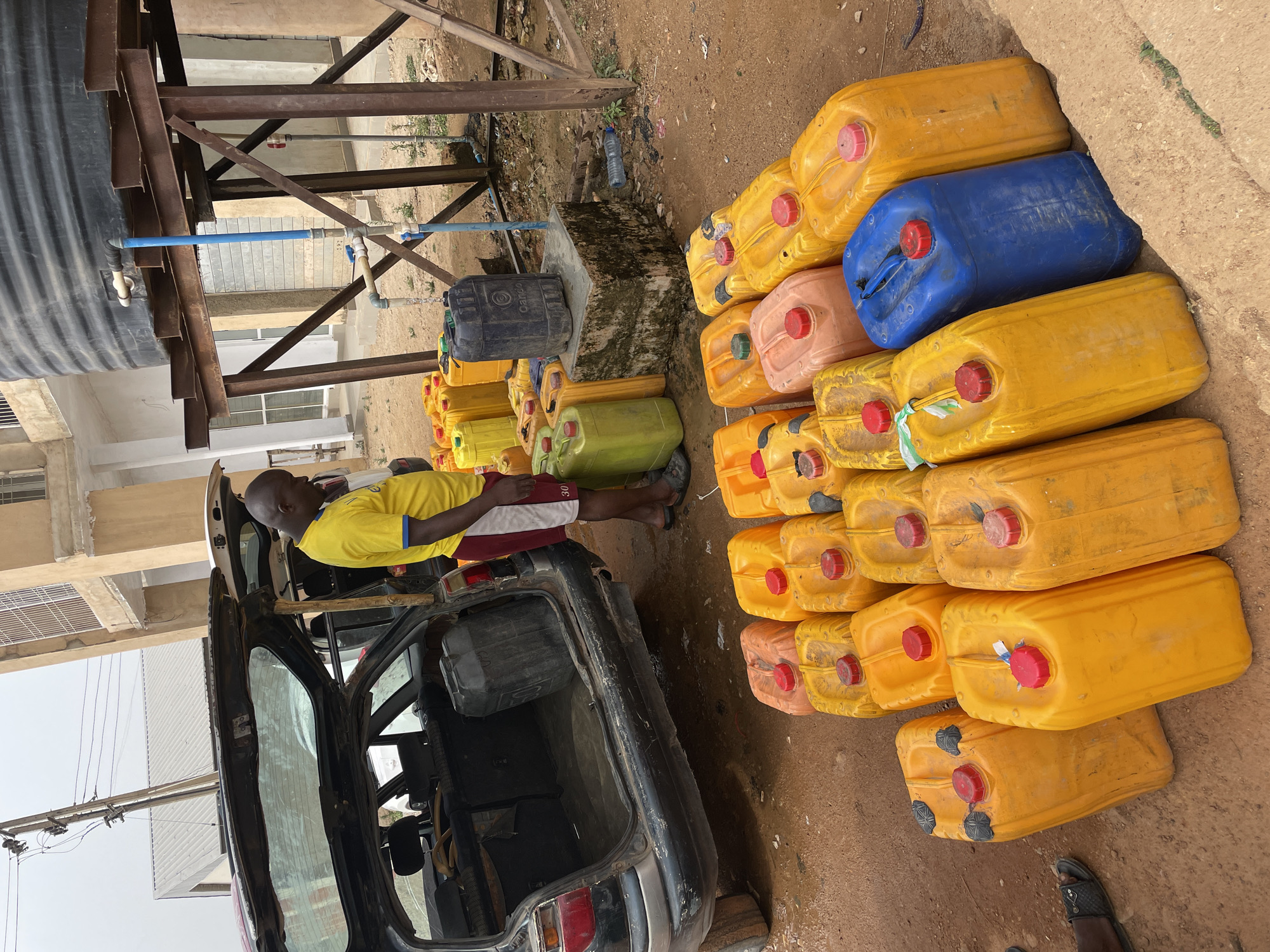 This image captures an outdoor scene, likely a water collection or distribution point, set on reddish-brown earth. In the foreground and midground, a large number of plastic jerrycans of various colors—predominantly yellow, but also orange, blue, green, and dark grey—are neatly stacked on the ground.
On the left side, a dark-colored car, possibly a hatchback or estate, has its boot open. A man with dark skin, wearing a yellow short-sleeved shirt and red and white shorts, is leaning over the open boot of the car, seemingly engaged in handling or preparing to load the jerrycans. His sandals are visible near the stacked containers.
Behind the car and to the left, a large, black plastic water tank rests elevated on a sturdy, rusty metal framework. Blue and white PVC pipes are connected to the tank, extending downwards with various fittings and valves, indicating a water delivery system. Below this structure, a concrete base holds another dark grey/black plastic drum.
The background reveals parts of light-colored buildings, possibly concrete structures, and overhead utility lines, suggesting an urban or semi-urban environment consistent with the provided location context of Jos, Nigeria. The overall lighting suggests it is daytime. No discernible text is visible on any objects. The scene depicts a common activity in regions where water is transported and stored in such containers.