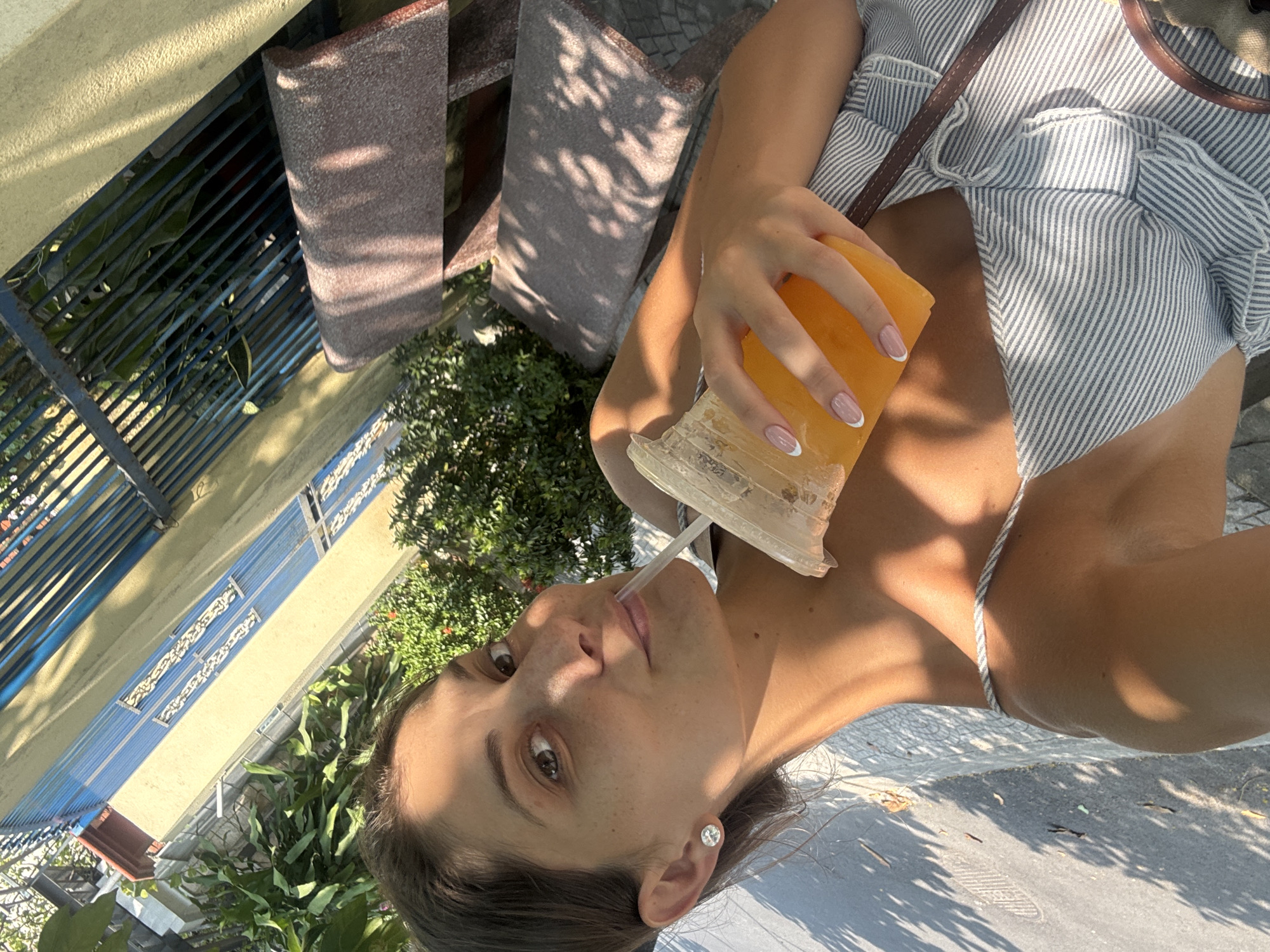 A young woman is holding a drink with a straw in a plastic cup. She is wearing a striped shirt and has a brown bag strap over her shoulder. She is wearing an earring in her left ear. The location is suggested to be Đà Nẵng, Vietnam, based on potential context that is not provided in the images. In the background, there is vegetation, a building with a blue railing, and outdoor seating with a patterned shadow.