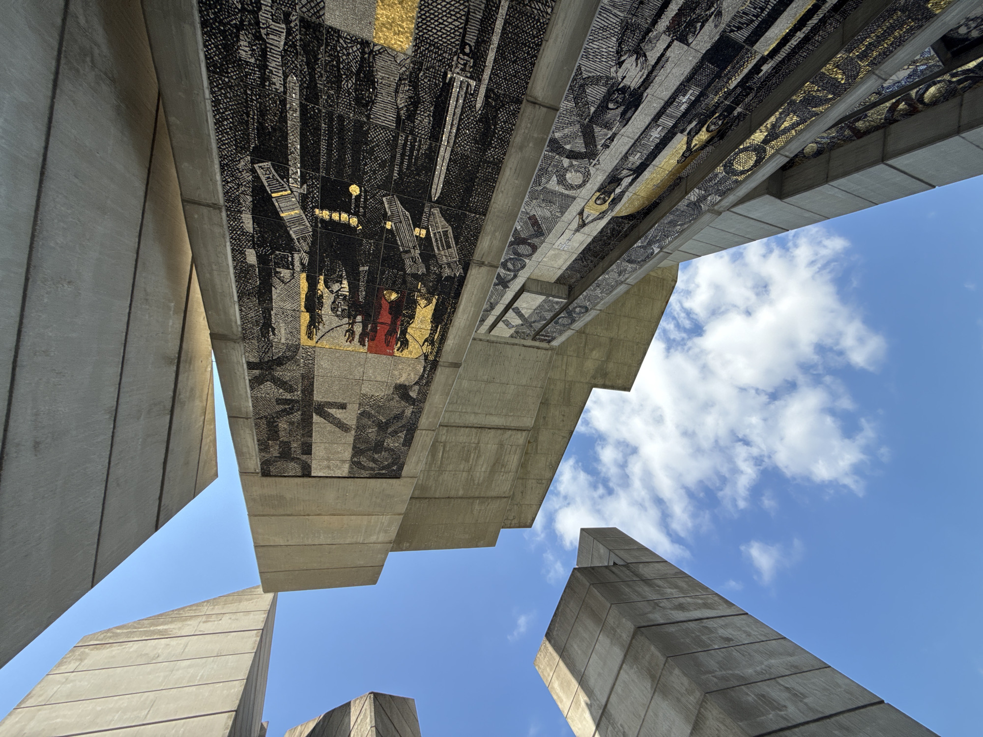 The image depicts a low-angle view of the Monument to the Declared Bulgarian State, also known as the Founders of the Bulgarian State Monument, located on a plateau near the city of Shumen, Bulgaria. The monument is constructed from concrete and features large, abstract geometric shapes forming towers and soaring structures. Integrated into the concrete surfaces are extensive mosaics.
The mosaics are intricate, primarily rendered in black, white, and gold tesserae, depicting historical scenes and figures. One mosaic prominently displays figures with weapons, possibly symbolizing historical battles or a military theme. Another mosaic shows a central figure with a golden halo, possibly representing a religious or spiritual leader, surrounded by other figures and symbols. The art style is consistent with Soviet-era socialist realist art, though the specific iconography here is tied to Bulgarian history.
The sky is a clear, bright blue with scattered white cumulus clouds, indicating a daytime scene. The lighting suggests the sun is high in the sky, casting some shadows on the concrete structures and highlighting the metallic elements of the mosaics. The perspective emphasizes the monumental scale and imposing architecture of the monument against the open sky. No people are visible in the frame. The environment is an outdoor setting, presumably on the plateau where the monument stands, offering an expansive view of the sky.