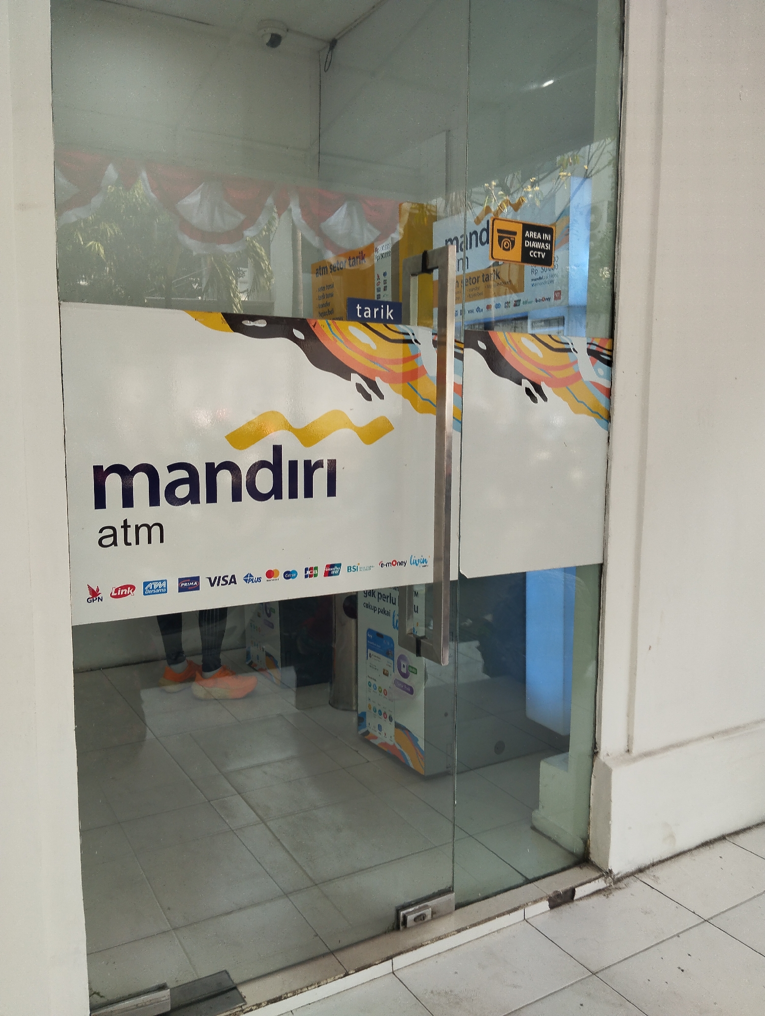 The image shows the entrance to a Mandiri ATM located in Surabaya, Indonesia. The ATM area is enclosed by glass doors. A prominent white banner with the Mandiri ATM logo and colorful abstract graphics is visible on the glass. Below the logo, various payment card logos are displayed, including GPN, Link, ATM Bersama, PRIMA, VISA, PLUS, Mastercard, JCB, BCA, BNI, BRI, Mandiri, and E-money Livin. A yellow sign indicating "AREA INI DIAWASI CCTV" (This area is monitored by CCTV) is also present on the glass. Inside the ATM area, reflections show red and white Indonesian flags and greenery outside, suggesting a festive or national occasion. The floor inside the ATM area is tiled. A person's legs, wearing orange shoes and dark pants, are visible through the glass, suggesting someone is inside the ATM area. A security camera is mounted on the ceiling above the entrance.