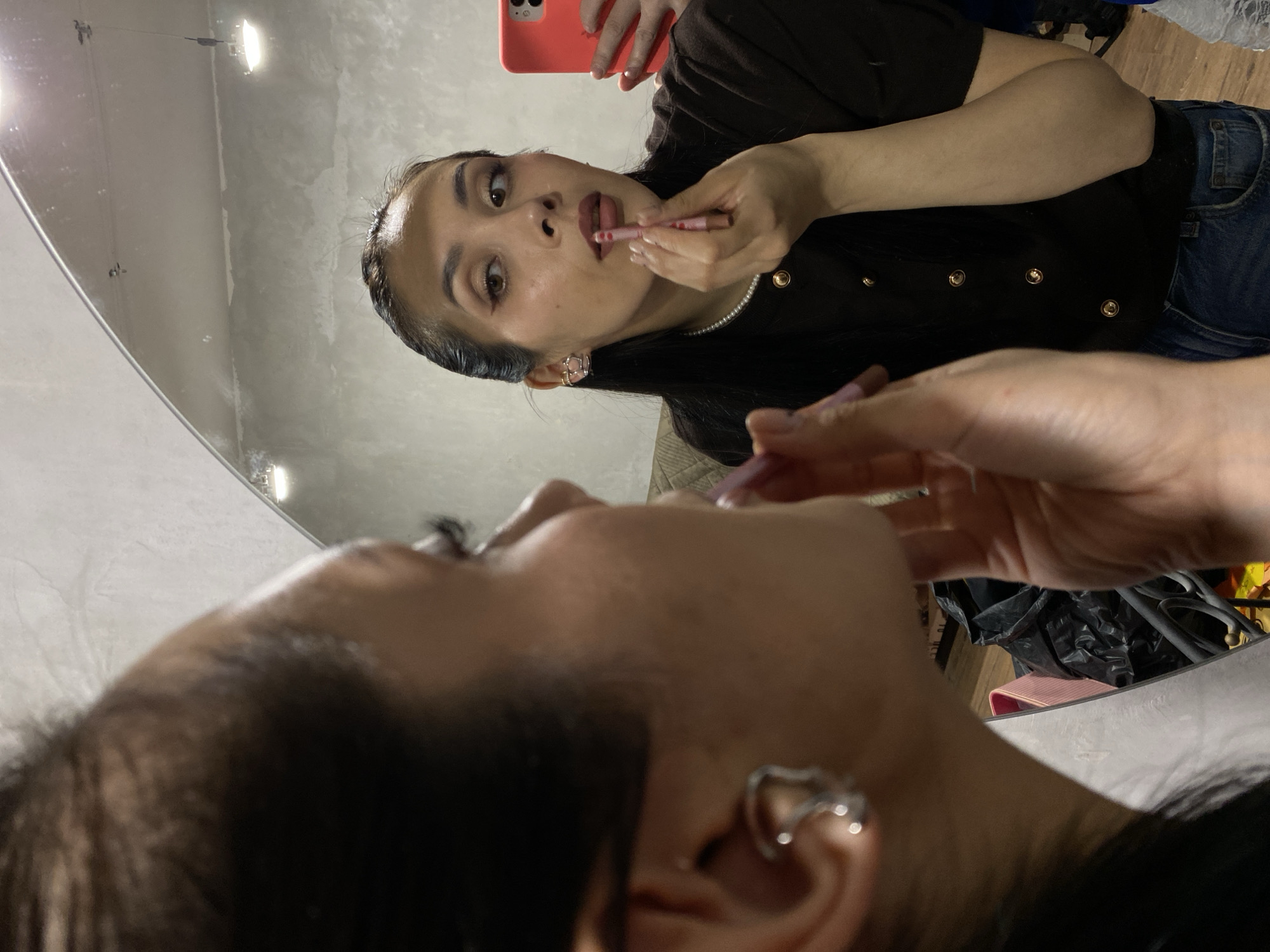 A young woman applies lipstick while looking at herself in a mirror. She holds a pink smartphone in one hand and a pink lipstick tube in the other. She wears a dark brown button-down shirt with a pearl necklace and blue jeans. The mirror reflects a close-up view of her face, revealing her earrings and makeup. The background is a gray wall with overhead lighting. There is no information to infer location.