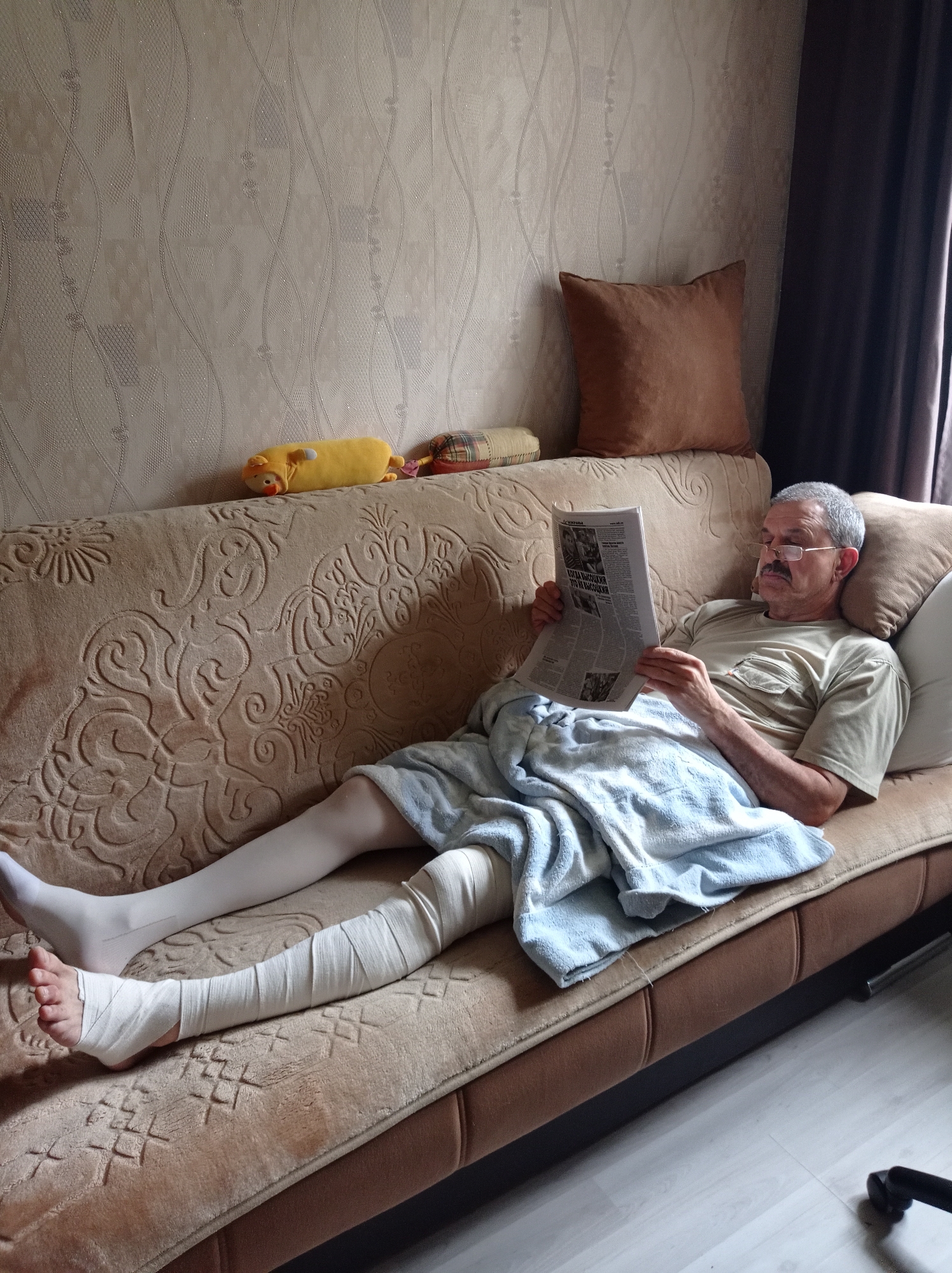 An elderly man with grey hair and a mustache, wearing glasses, a light-colored t-shirt, and compression stockings, is sitting on a couch reading a newspaper. His right leg is bandaged from the ankle to the knee, and his left leg is also in a compression stocking. A light blue towel is draped over his legs. He is in a room with patterned wallpaper and a brown couch with decorative stitching. A brown pillow is behind his head, and another pillow is to his right. Two plush toys, a yellow pig and a checkered object, are on the couch behind him. The scene appears to be indoors, likely in a residential setting in the city of Degtyarsk, Russia.