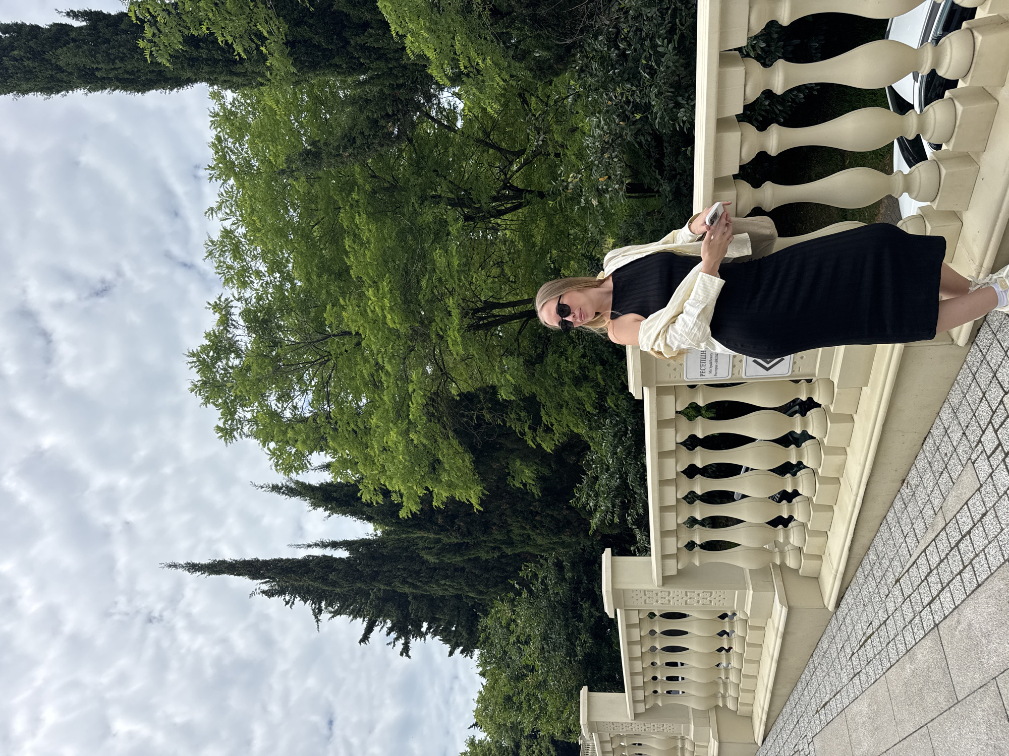 A young woman wearing sunglasses, a black dress, and a light-colored jacket stands on a paved walkway next to a beige stone balustrade. She is holding a smartphone in her right hand and a small bag is slung over her left shoulder. Behind her, a dense green forest with tall cypress trees and other deciduous trees fills the background, under a cloudy sky. The scene appears to be located in Sochi, Russia, potentially in a park or garden area with an ornate railing.