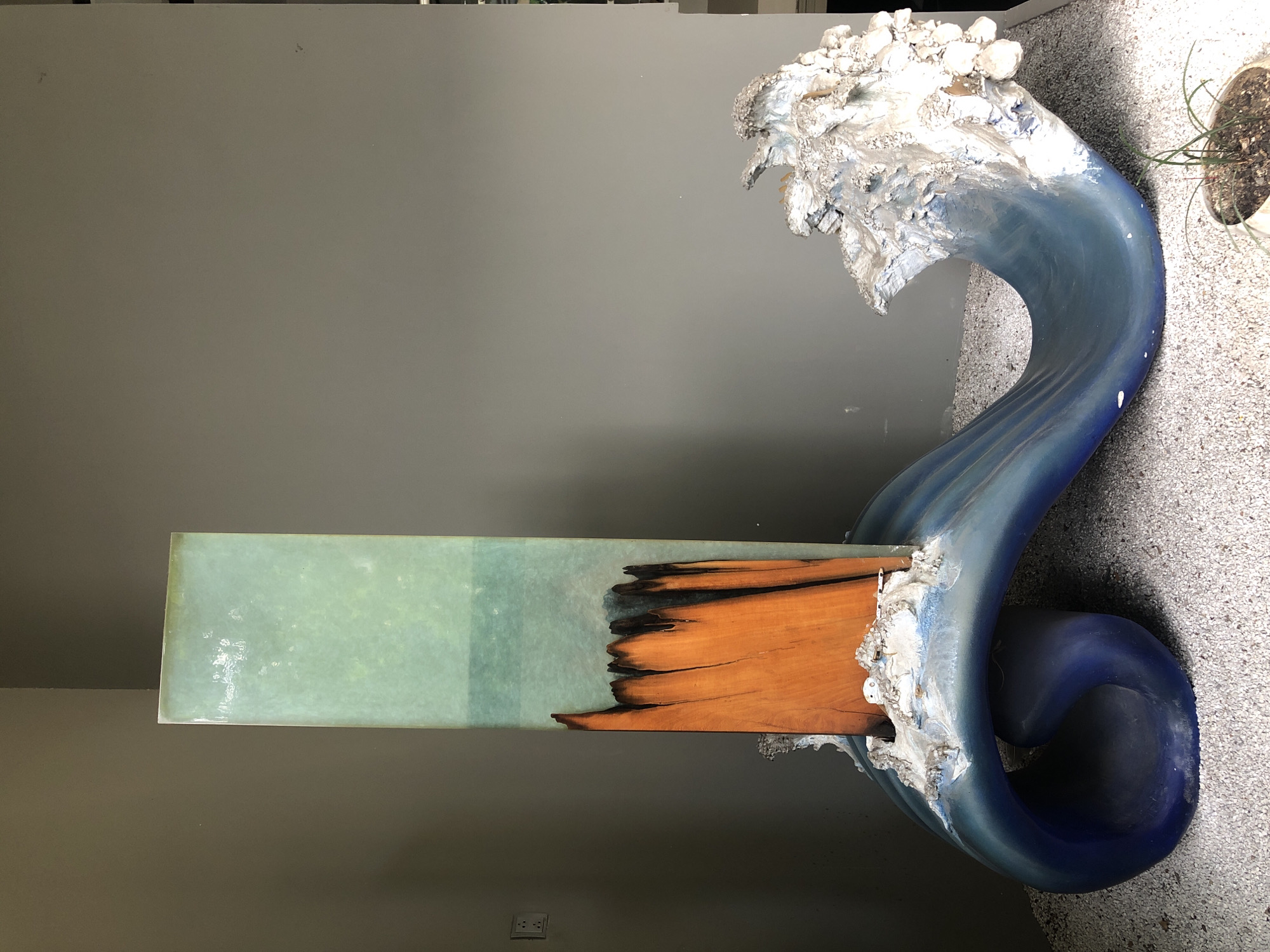 This image captures a striking sculpture situated indoors against a plain, grey wall. The sculpture features a tall, rectangular, translucent green element on the left, with a vibrant orange wooden piece embedded within its lower section. This wooden piece has a rough, jagged top edge and appears to be partially submerged in white, foamy material. The sculpture rests on a large, dark blue wave-like base that curves and curls, with white foam cresting on the right side. The base is supported by a bed of small, light-colored pebbles. A small potted plant with green shoots is visible in the lower right corner. The overall impression is one of a dynamic seascape frozen in time, with the sculpture likely representing a stylized wave crashing against an abstract form. There is no discernible indication of its specific location within the city of Đà Nẵng, Vietnam.