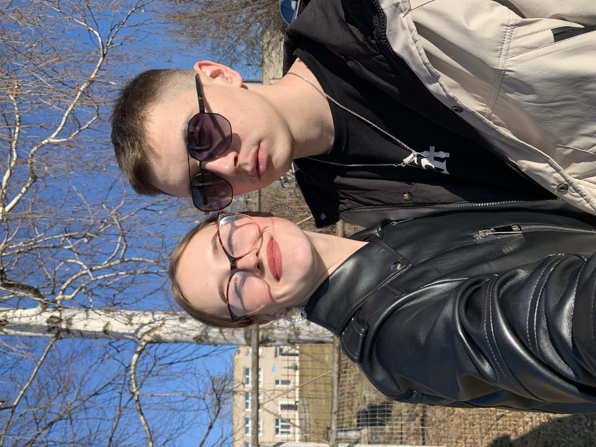 A portrait-oriented photo captures two young individuals, a male and a female, posing outdoors under a clear blue sky. The male, positioned in the upper left of the frame, has short dark hair and wears sunglasses resting on his nose. He has a neutral expression and is dressed in a light-colored, possibly beige, windbreaker jacket over a black shirt with a silver chain necklace featuring a prominent cross pendant.
The female, in the lower right, wears eyeglasses with patterned frames and has light brown hair. She displays a slight pout or playful expression with pink lipstick and is wearing a black leather jacket over a dark top, accessorized with a small pearl necklace.
The background features a bright blue sky with numerous bare tree branches, some displaying the white bark characteristic of birch trees. In the distance, a light-colored, multi-story building with windows is visible, suggesting an urban or suburban environment. The bright sunlight indicates a clear day, possibly late winter or early spring, consistent with the location of Фоки, Russia. No visible text is present in the image.