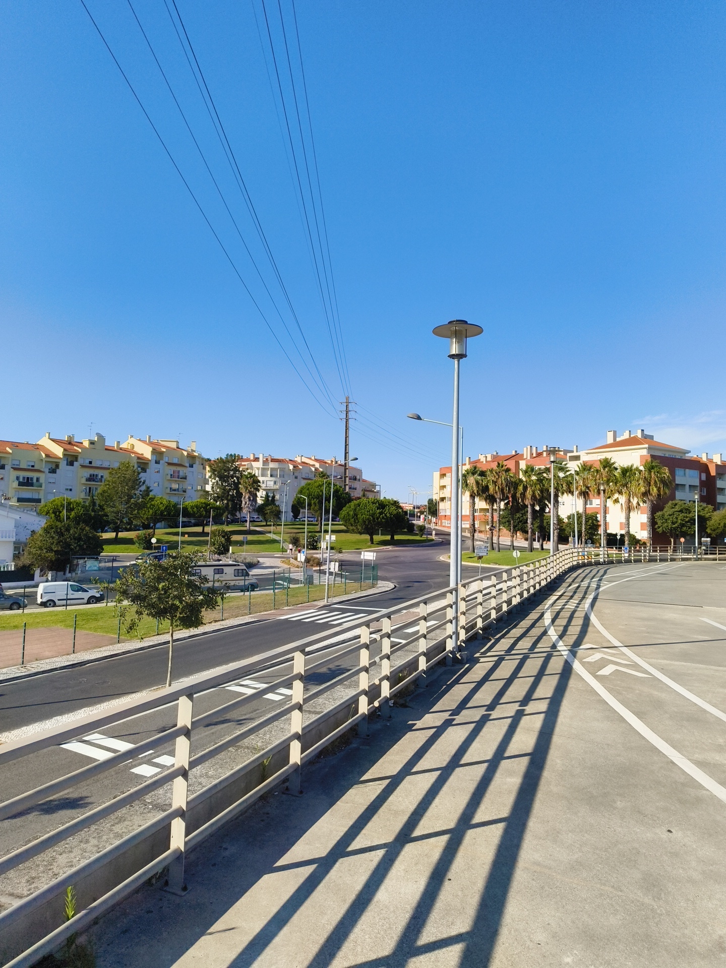 The image depicts a view of a pedestrian walkway and street in Cascais, Portugal. The walkway features a light-colored metal railing casting long, parallel shadows across the concrete surface. Beyond the walkway, a road curves to the left, with a pedestrian crossing marked by white stripes. On the other side of the road, a grassy embankment is visible, leading to a row of apartment buildings with orange-tiled roofs. Trees and palm trees are interspersed among the buildings and along the embankment. Overhead, power lines stretch across the clear blue sky. The scene appears to be in a residential area, likely on a sunny day. No people are present in the image.