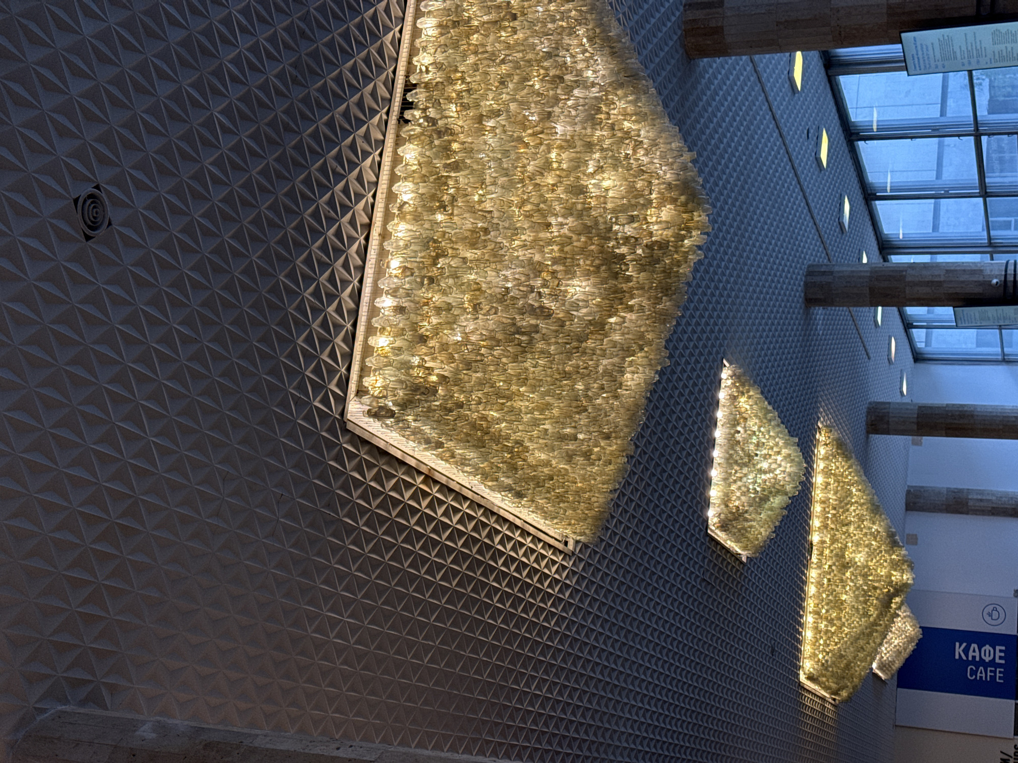 The image shows an interior space in Moscow, Russia, featuring a textured wall with geometric patterns. Three rectangular illuminated chandeliers hang on the wall, constructed from hundreds of small, light-colored elements. A sign with the text "КАФЕ CAFE" indicates a cafe or dining area. The space also features a window towards the top of the image, providing natural light and a view of the outside. There are structural beams running horizontally across the top of the wall.