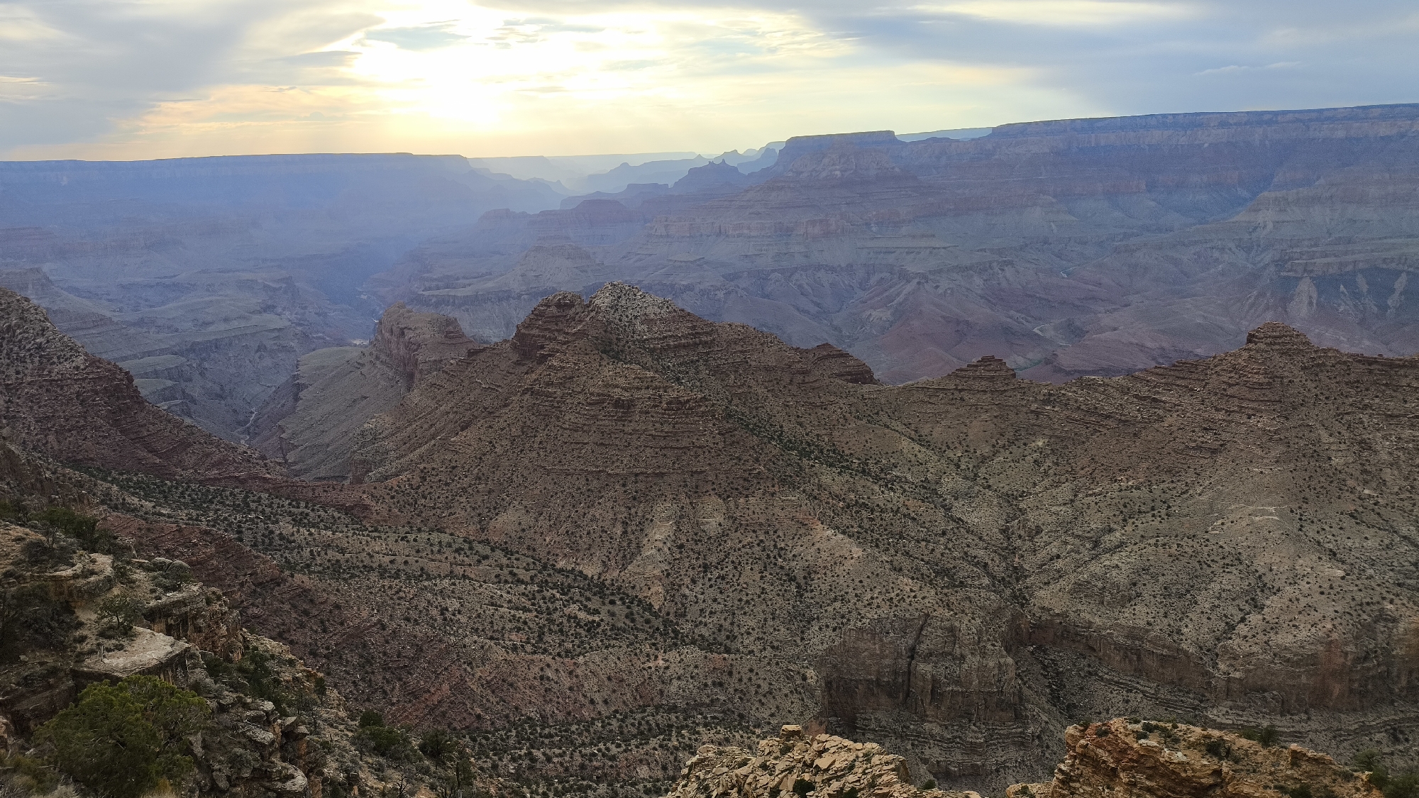The image depicts a wide, layered landscape of the Grand Canyon, viewed from a high vantage point. The foreground is dominated by rugged, rocky terrain with sparse vegetation. Beyond this, the canyon opens up into a vast expanse of sculpted rock formations, displaying various shades of red, brown, and gray, indicative of different geological strata. The layers of the canyon recede into the distance, becoming progressively lighter and more muted due to atmospheric haze.
The sky above is partly cloudy, with the sun breaking through the clouds in the upper left portion of the frame, casting a soft, diffused light over the scene. The light creates a sense of depth and highlights the contours of the canyon walls. The time of day appears to be late afternoon or early evening, suggested by the angle of the sunlight and the soft, diffused lighting.
The scene is devoid of any people or man-made structures, presenting a natural, untouched panorama. The environment is characteristic of the arid, elevated plateau of the Grand Canyon National Park, located in Arizona, United States. The immense scale of the geological formations suggests a location within the Grand Canyon Village area, a common viewpoint for observing the vastness of the canyon.