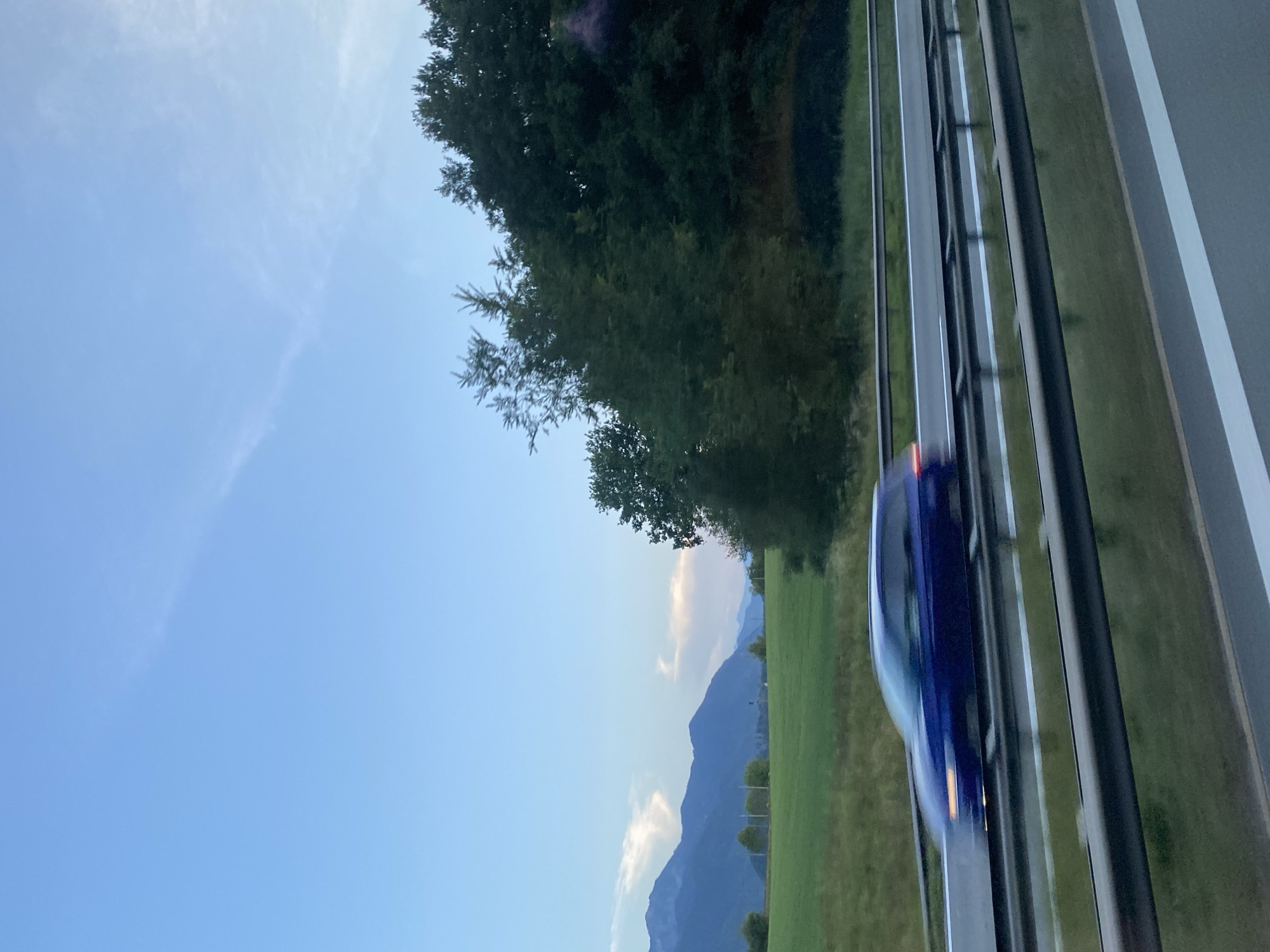The image captures a highway scene near Grabenstätt, Germany, during what appears to be late afternoon or early evening. A dark blue car, blurred by motion, travels on the highway in the foreground. The highway has multiple lanes separated by white lines and guarded by metal barriers. Beyond the highway, a lush green field stretches towards a dense line of trees. In the background, a series of majestic mountains form the horizon under a pale blue sky with wispy clouds. The overall impression is one of movement and a scenic rural landscape.