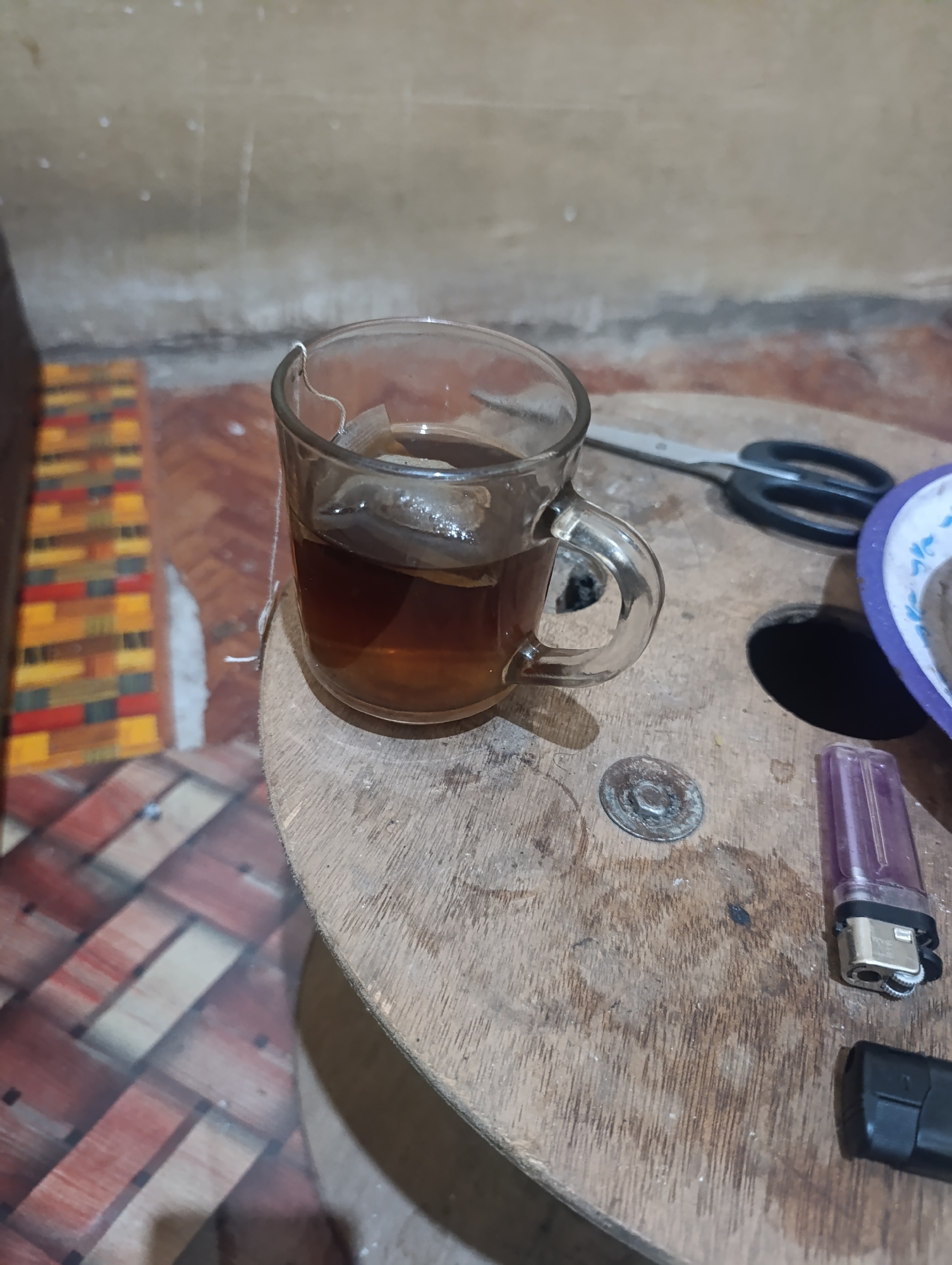 A close-up, eye-level shot displays a clear glass mug filled with dark tea and a tea bag steeping inside, positioned on a weathered wooden surface. The mug's handle is visible on the right side of the glass. To the left of the mug, a colorful, woven mat is partially visible, featuring red, yellow, and black rectangular patterns. Behind the mug, a pair of black-handled scissors with silver blades rests on the wooden surface. Further to the right, a hole is present in the wooden surface, and a purple plastic lighter and a black object, possibly another lighter, are also on the wood. A purple and white plate with a floral pattern is located on the far right. The background consists of a textured, neutral-colored wall with faint speckles. The scene is lit, casting subtle shadows. This scene is located on a wooden spool table in Madatte, Indonesia.