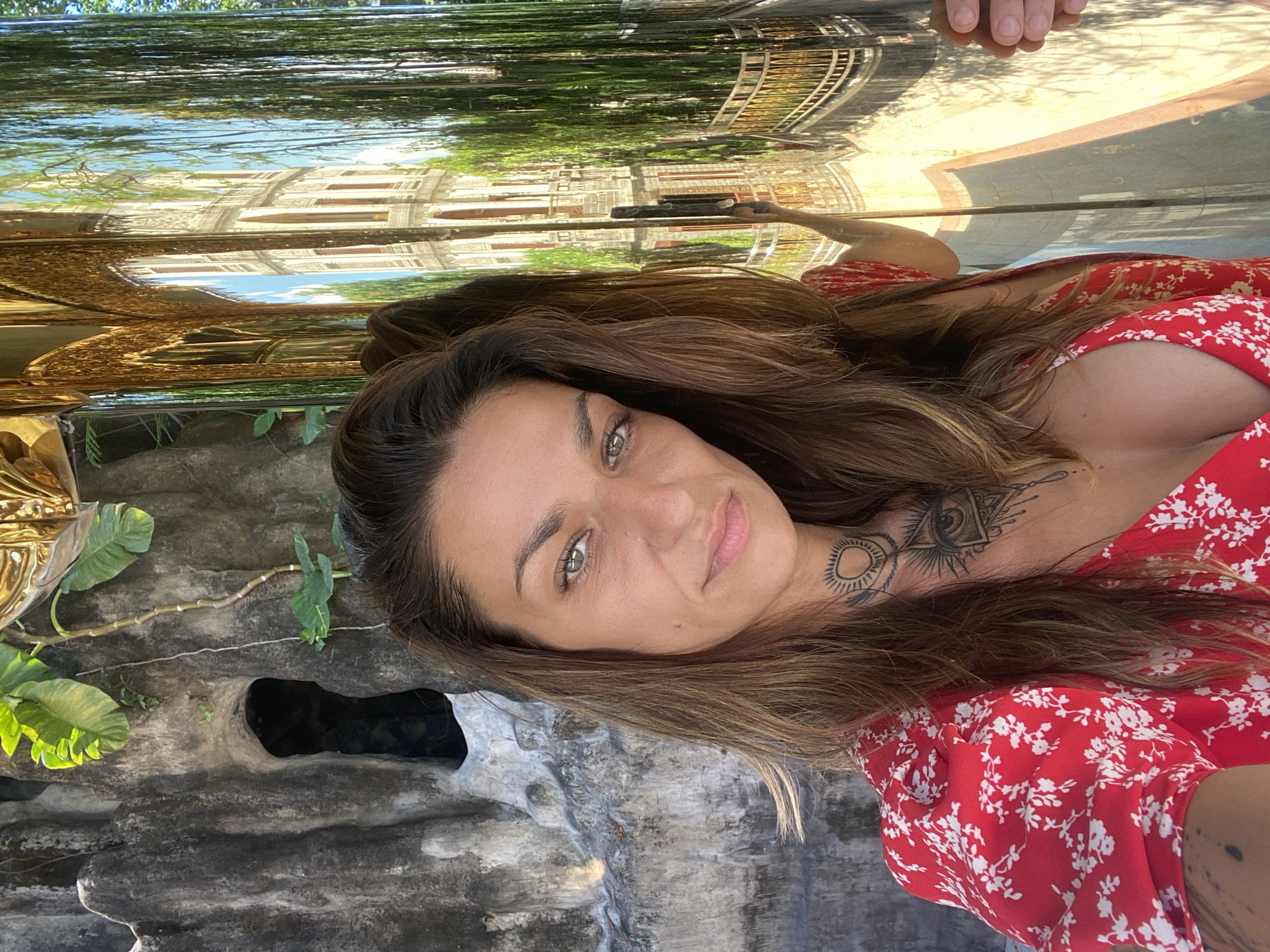 A woman with brown hair and fair skin is posing for a selfie outdoors. She is wearing a red dress with white floral patterns. She has tattoos on her neck and arm. The background includes a stone-like structure with foliage, and a reflective surface above shows a partial reflection of buildings and trees, possibly in a city environment. The location is likely Dĩ An, Vietnam, as requested. A hand is seen in the reflection above her head, possibly holding the phone.