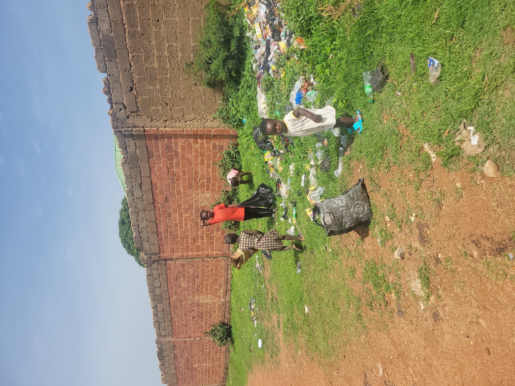 In Jos, Nigeria, a group of children are observed near a brick wall bordering an area strewn with garbage. Three children are visible in the foreground: two are standing, facing away from the camera, and one is a toddler. The child in the orange shirt is holding a black trash bag, while the other child, wearing a plaid dress, holds a brown paper bag, possibly for collecting refuse. The toddler, in a light-colored shirt and blue sandals, stands facing the camera. The environment is characterized by overgrown grass, littered with various types of trash including plastic bottles, bags, and wrappers. Beyond the children, a larger pile of refuse is visible, partially obscured by dense green vegetation. The background features a partially constructed brick wall. The sky is clear and blue.