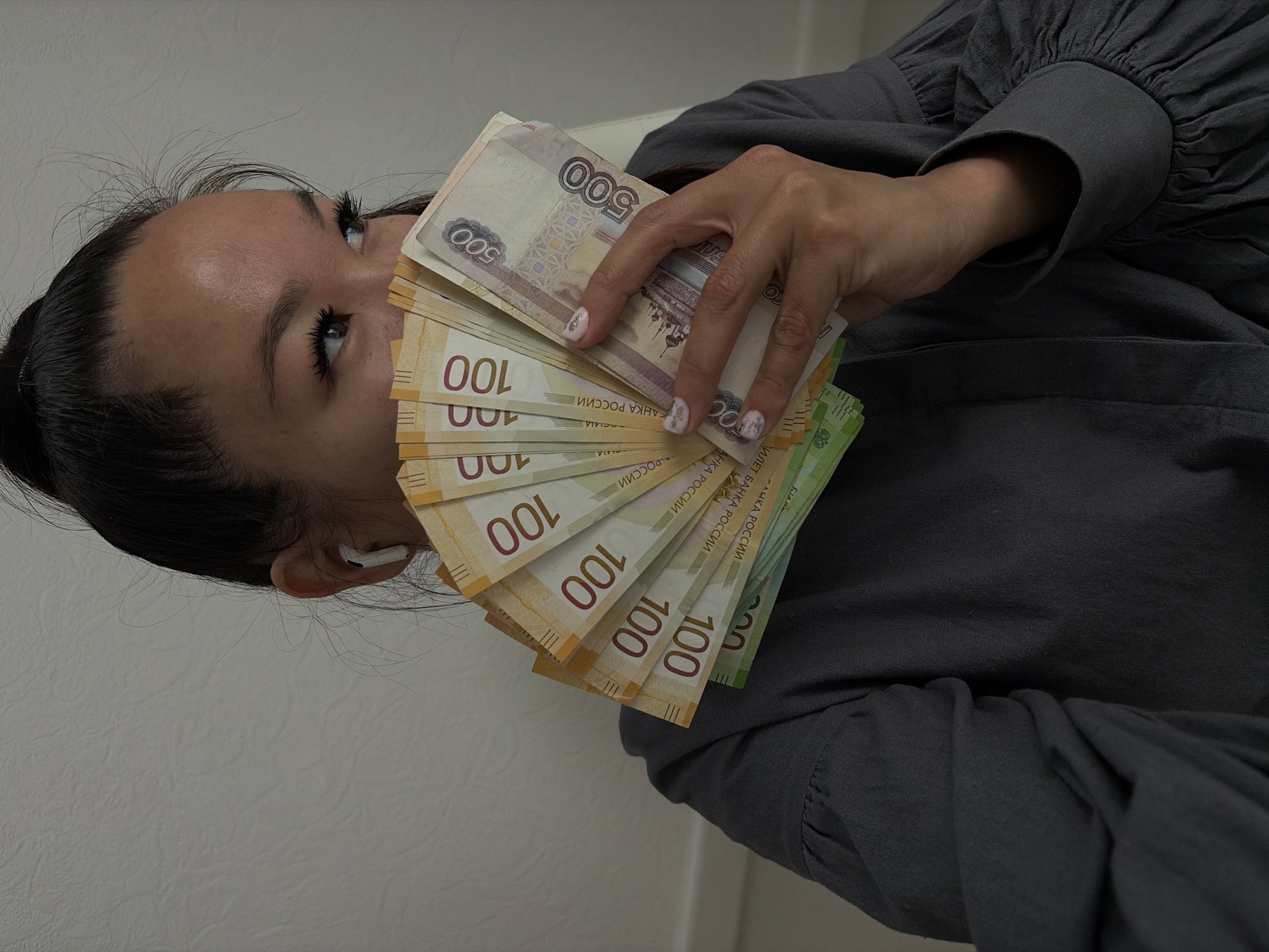In a medium shot, a woman is partially visible, holding a fan of Russian rubles in front of her face. She has dark hair pulled back into a bun and is wearing white earbuds. Her eyes are looking to her right, and she has dark mascara on her eyelashes. Her fingernails are painted with a light-colored polish and have small floral designs. The background appears to be a textured, light-colored wall. The woman is wearing a dark gray blouse with puffed sleeves. The money held by the woman consists of several 100-ruble notes and at least two 500-ruble notes. There is no visible indication of the city of Chaikovsky, Russia, within the image.
