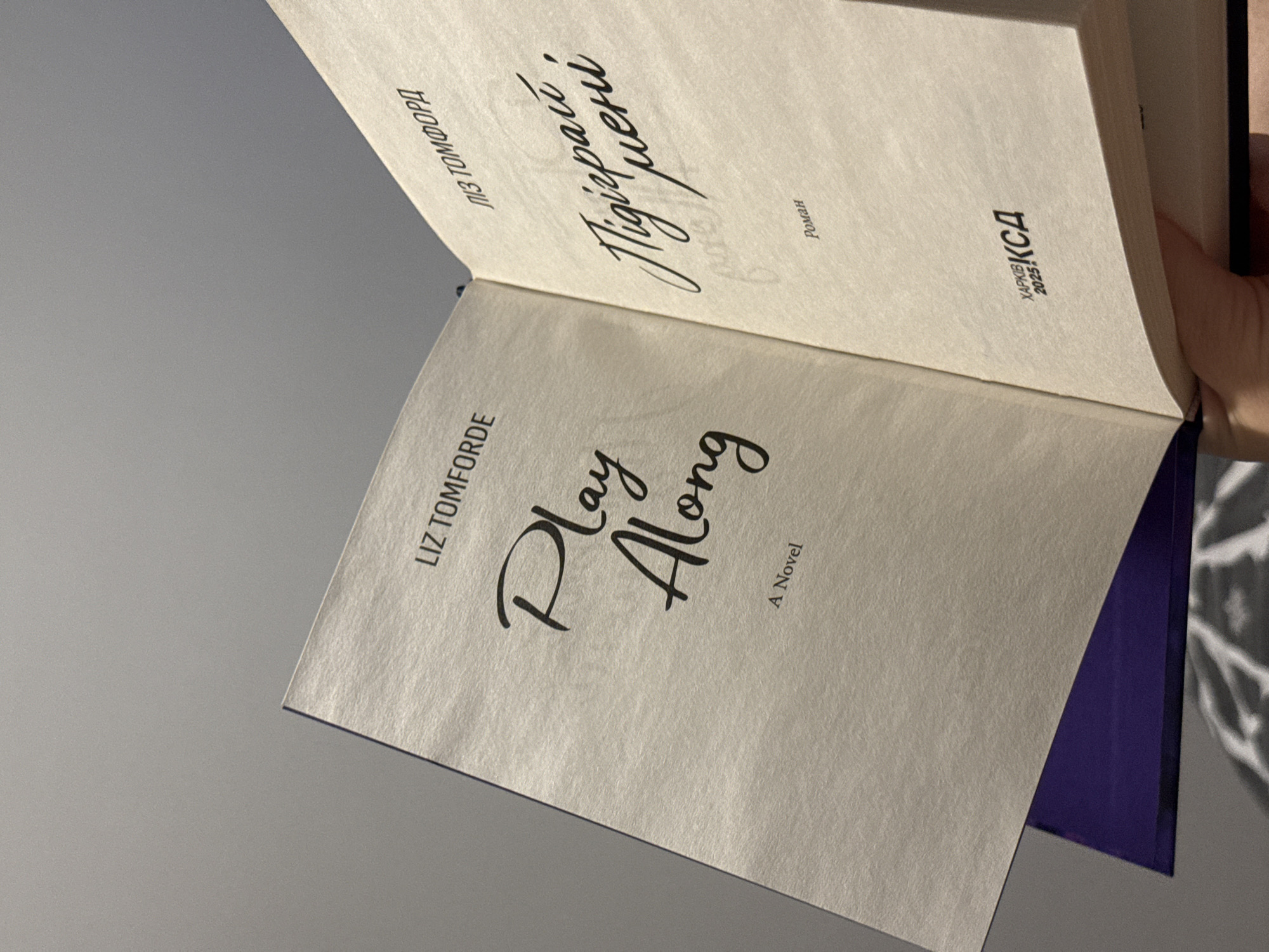 This image shows an open book held by a person's hand. The left page of the book displays the title "Play Along" in a large, stylized font, with "A Novel" printed below it in a smaller font. Above the title, "LIZ TOMFORDE" is written. The right page shows text in a script font, which appears to be in a different language, possibly Ukrainian, with "ЛІЗ ТОМФОРД" above it. Below the script text, "Роман" is printed, and at the bottom of the page, there is a logo with the text "ХАРКІВ 2025 КСД". The background is a plain grey surface, and a purple object is visible beneath the book. There is no discernible connection to the city of Łódź, Poland.