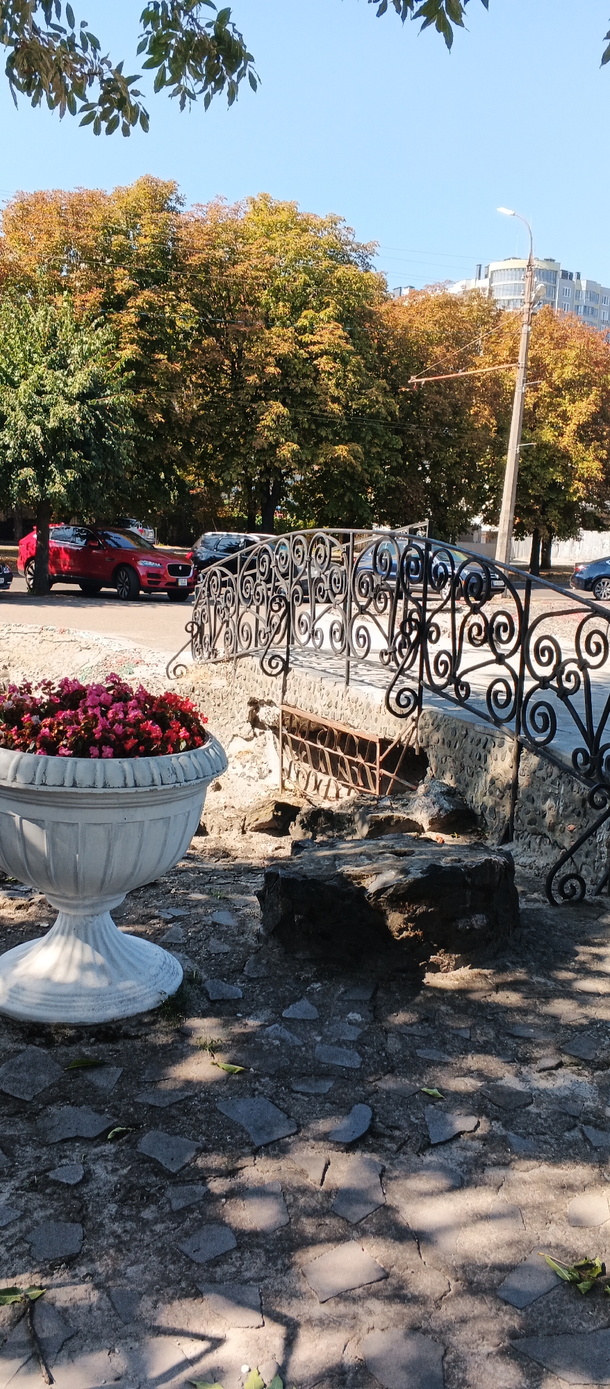 This image depicts a street scene in the city of Cherkasy, Ukraine. In the foreground, a large, ornate white planter overflows with vibrant pink and red flowers. To the right of the planter, a section of a decorative wrought-iron fence with scrolled accents runs along what appears to be a small, stone-lined bridge or walkway. The bridge itself is partially visible, constructed from rough stones and featuring a metal grate underneath. A large, dark rock sits on the paved ground in front of the bridge.
The ground is paved with irregular stone tiles, and shadows from tree foliage are cast across it. Behind the planter and fence, a red SUV is parked on the side of a street lined with trees. These trees display the warm hues of autumn, with leaves transitioning from green to yellow and brown. Further in the background, several other vehicles are visible on the street, and a modern, multi-story building with balconies stands against a clear blue sky. A lamppost with a light fixture is also present. The scene is captured on a sunny day, indicated by the bright lighting and distinct shadows.
