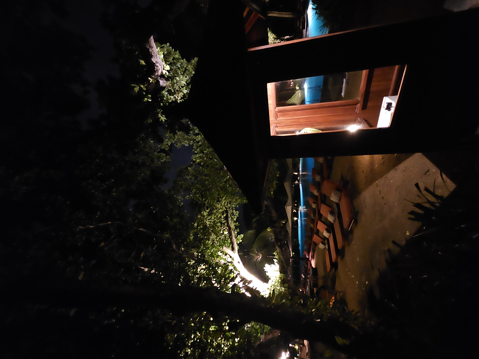 The image depicts an outdoor scene at night in Denpasar, Indonesia. A building with warm interior lighting is visible through a window, contrasting with the darkness of the surrounding environment. Adjacent to the building is a swimming pool illuminated by blue light. Rows of seating, possibly lounge chairs, line the pool area. The scene includes foliage such as trees and plants, some illuminated by spotlights or other light sources, adding depth and texture to the image. The perspective is skewed, as the camera seems tilted.