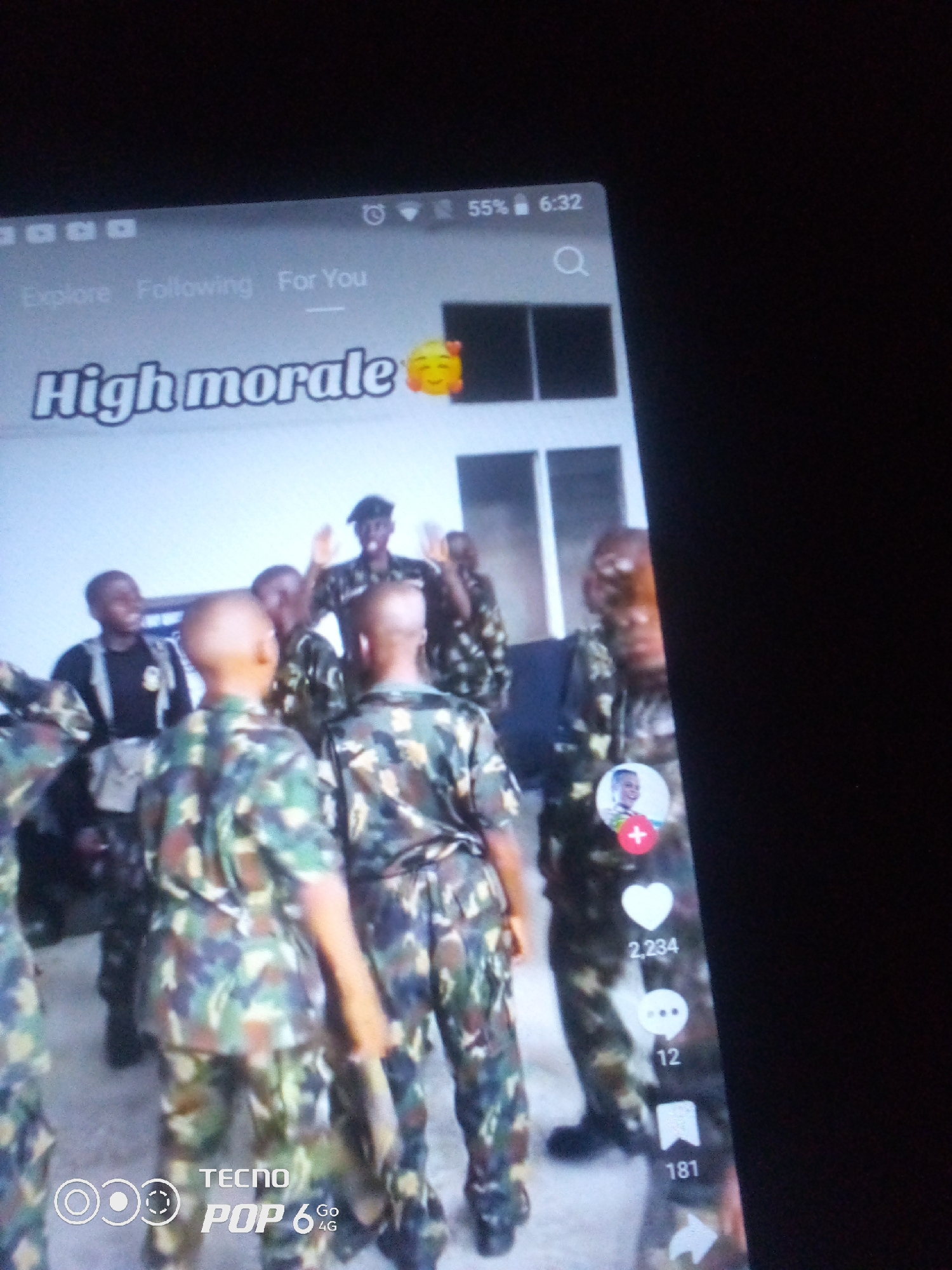 The image is a vertical, slightly blurry photograph taken on a mobile device, likely a Tecno Pop 6 4G, as indicated by the watermark at the bottom left. The screen displays a social media interface, possibly TikTok, featuring a video with several individuals in camouflage military uniforms. The video's caption reads "High morale" followed by a smiling face emoji. Above the caption, navigation tabs "Explore", "Following", and "For You" are visible. The top right corner of the screen shows the battery status as 55% and the time as 6:32. Social media interaction icons, including a heart with "2,234" likes, a speech bubble with "12" comments, and a bookmark with "181" shares, are arranged vertically on the right side of the screen. A circular profile picture with a red plus sign is also visible within the social media interface. The background of the image is dark, suggesting the photo was taken in low light conditions, possibly at night or indoors. There is no indication of the city of Howayi, Nigeria, or any other specific geographical location within the image itself. The image primarily captures a digital display of a social media post.