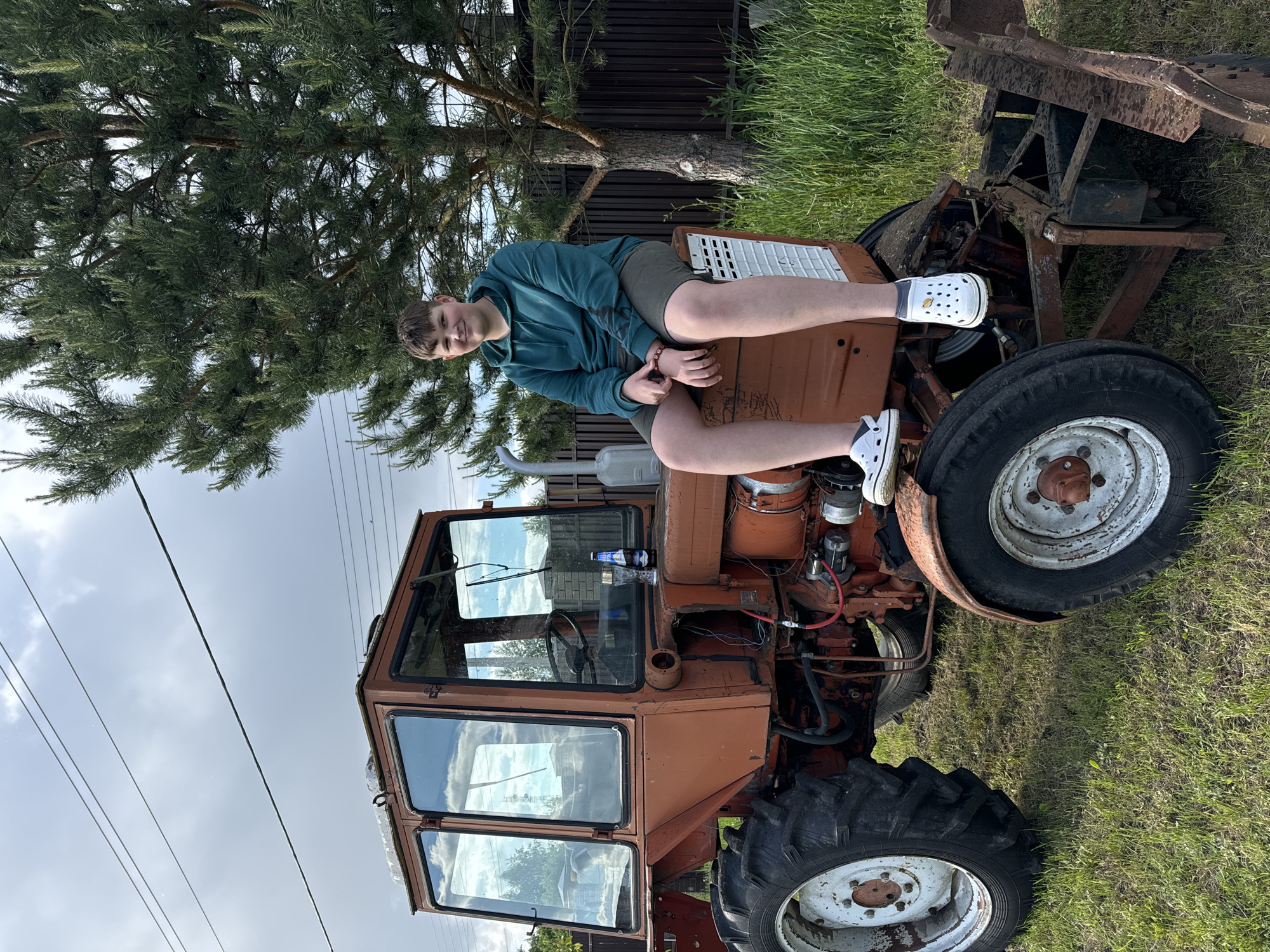 A young person is reclining on the hood of an orange tractor, positioned on a grassy field. The tractor, partially obstructed by a tree, has a cabin and large tires. The background includes a fence and a cloudy sky with overhead power lines. The specific location is Vysokinichi, Russia.