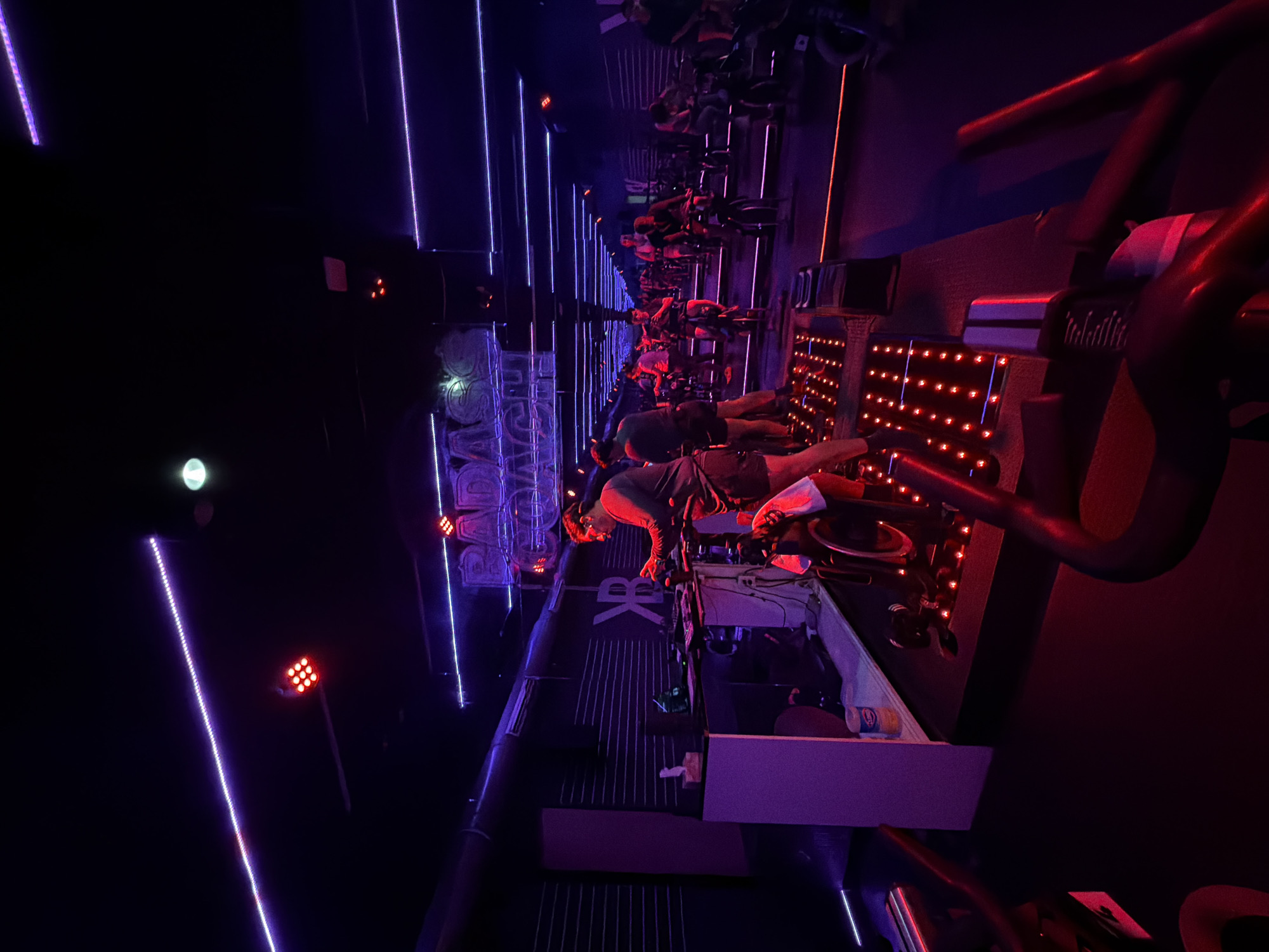The image depicts an indoor cycling studio, likely in Puebla, Mexico, given the "BADASS" signage. The room is illuminated with a combination of purple and orange LED lighting. Rows of stationary bikes are visible, many with individuals participating in a class. The perspective suggests the image was taken from one of the bikes, looking towards the front of the studio.