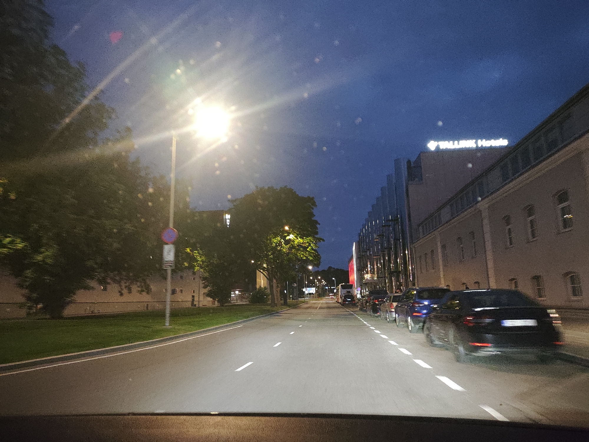 The image depicts a nighttime street scene in Tallinn, Estonia. A street lamp illuminates the road, casting a bright light and creating lens flare. Tall trees line the left side of the road, with a grassy area and a building visible behind them. A street sign with a red and blue circular symbol is present. On the right side of the road, a modern building with glass windows and vertical elements stands, with a sign that reads "TALLINK Hotels" illuminated at the top. Several cars are parked and driving on the right side of the road, including a black sedan in the foreground. The sky is dark blue, indicating it is dusk or night. The perspective suggests the image was taken from inside a vehicle.