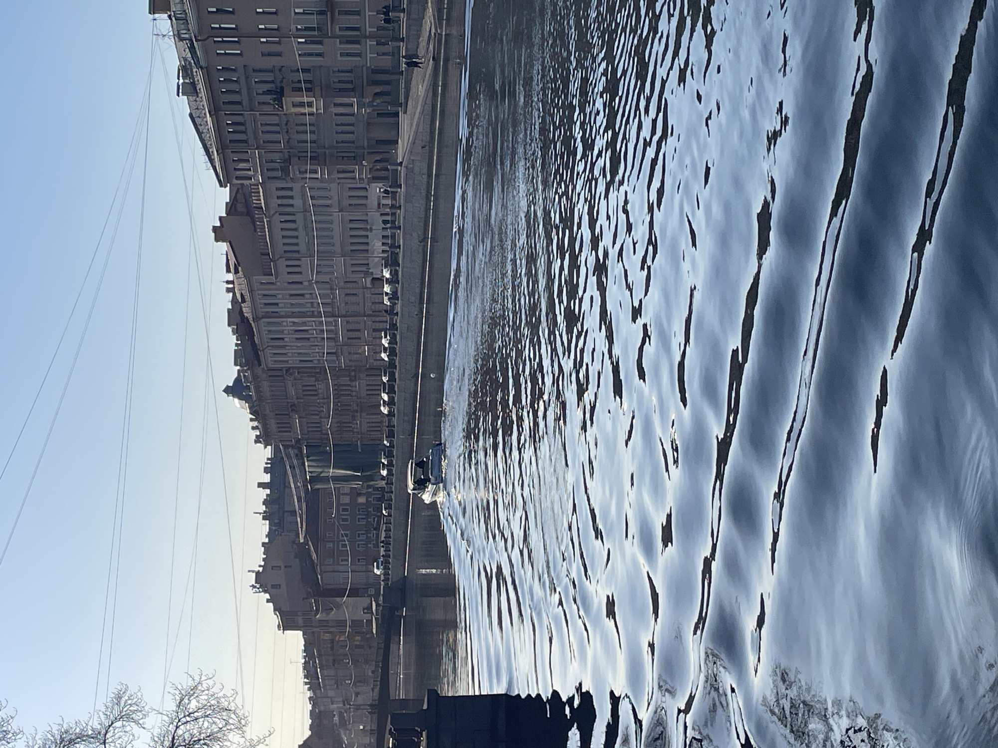 This image depicts a sunlit urban canal scene on the Fontanka River in Saint Petersburg, Russia, likely during late winter or early spring due to bare tree branches in the upper left. A small, white motorboat with a dark hull is prominently featured in the middle distance, traveling away from the viewer and generating a distinct V-shaped wake across the water's surface. The canal water reflects the bright sky and exhibits numerous ripples from the boat's movement and natural currents.
On the right side of the waterway, a stone embankment with a wide paved pedestrian area runs parallel to the canal. Several indistinct individuals are visible walking along this sidewalk. Adjacent to the pedestrian area is a street lined with approximately ten to twelve parked automobiles. Beyond the street, a continuous row of multi-story historic buildings with light-colored facades extends into the distance. These buildings feature numerous windows, ornate architectural details, and multiple chimneys. One building facade further down the embankment is partially covered by green netting, suggesting ongoing renovation or construction.
In the background, a multi-arched stone bridge crosses the canal, with additional historic buildings visible on the far side. The sky is clear and bright blue, traversed by several thin utility wires overhead. The left side of the foreground is partially obscured by dark, out-of-focus stone structures and the aforementioned bare tree branches. The scene is illuminated by strong sunlight, casting reflections on the water and highlighting the building facades.