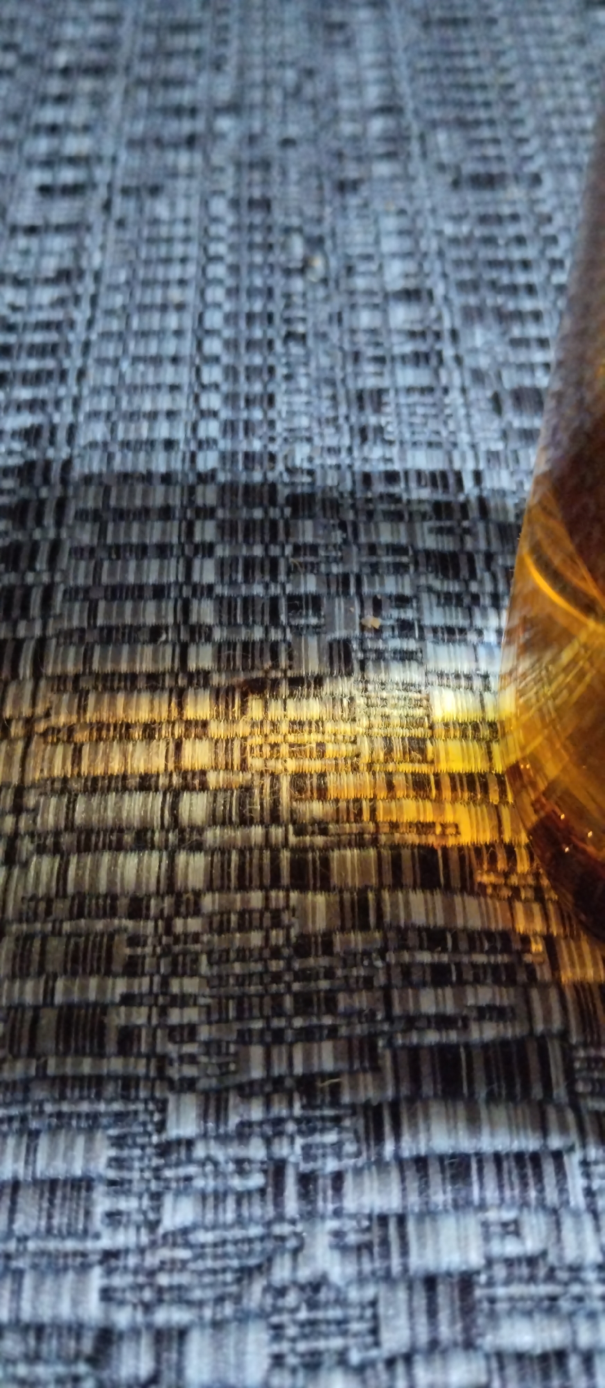 The image shows a close-up view of a woven textile with a pattern of horizontal and vertical lines in shades of gray, black, and white. A shaft of golden light falls diagonally across the fabric, illuminating a section of it. On the right side of the image, a portion of a glass bottle with amber-colored liquid is visible. The bottle appears to be reflecting the light. The texture of the fabric is rough and tactile. The scene is indoors, possibly on a table or surface covered with this textile. There are no people or animals visible in the image. There is no information in the image that would suggest a location in the city of Sondrio, Italy.