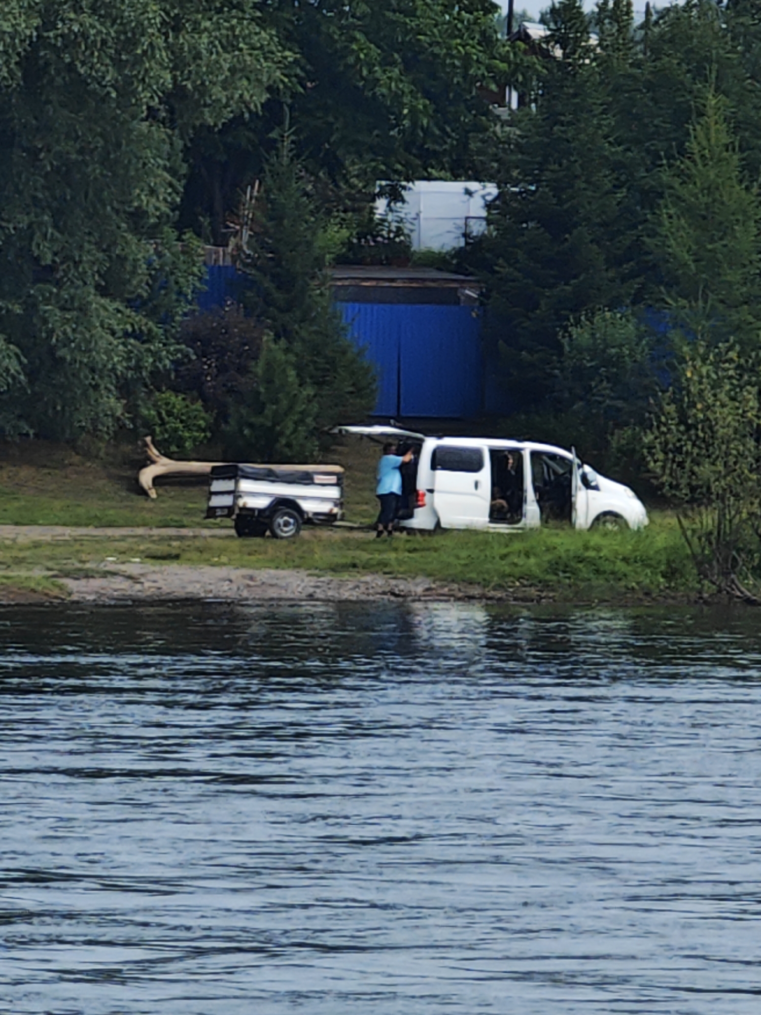 In Sosnovoborsk, Russia, on the shore of a body of water, a white minivan with its rear hatch and passenger side door open is parked on a grassy area. A person wearing a blue top is standing by the open rear hatch, and another person is visible in the driver's seat. Adjacent to the minivan, a small white trailer is parked. In the background, a blue fence and trees are visible. A large, gnarled tree trunk lies on the ground near the trailer. The water in the foreground appears calm with ripples.