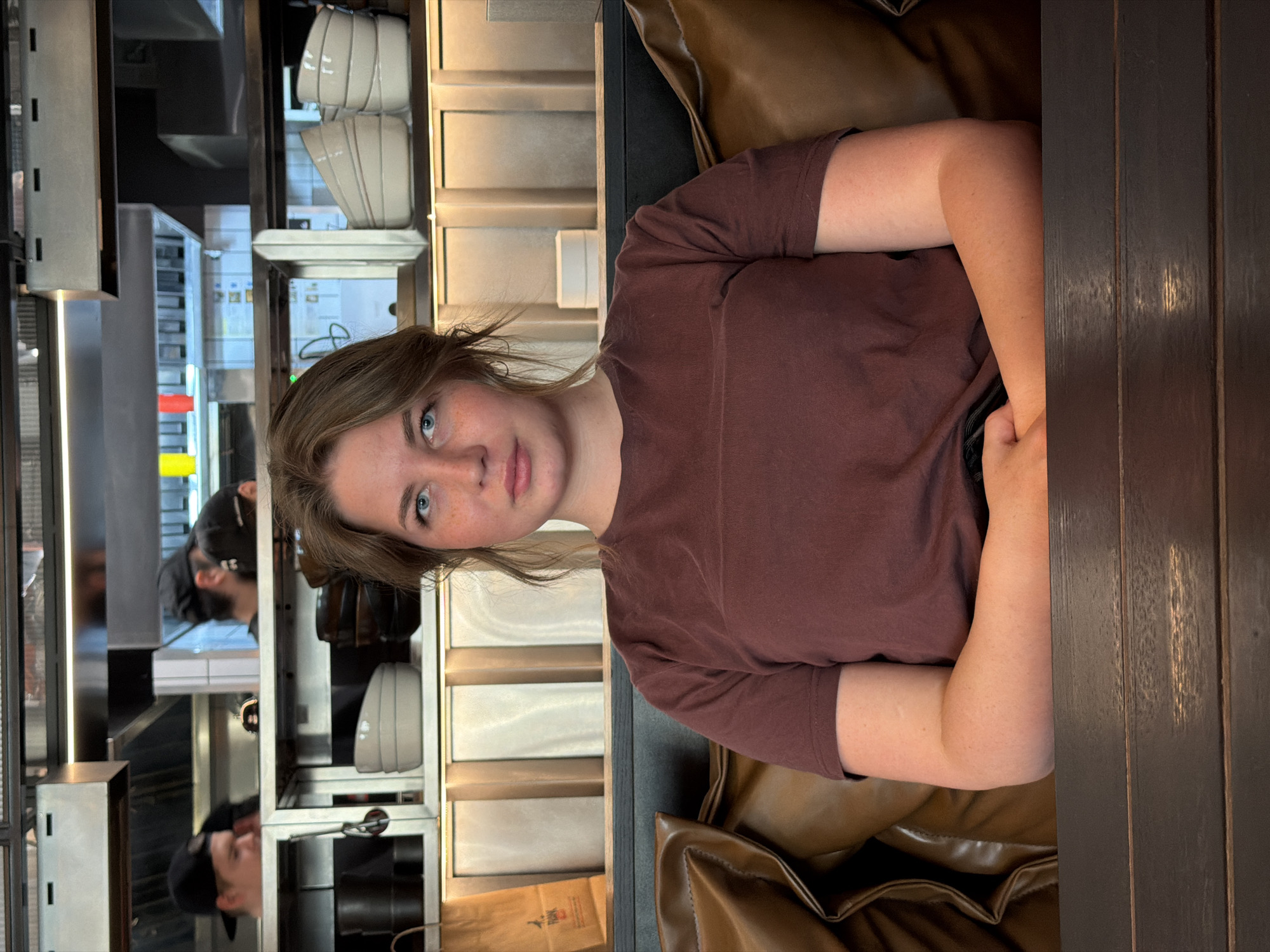 A young adult female is seated at a dark wooden table in a casual dining establishment in Kazan, Russia, looking directly at the camera with a neutral expression. She has medium-length light brown hair, blue eyes, and light freckles on her nose and cheeks. She is wearing a plain, short-sleeved, reddish-brown t-shirt. Her forearms rest on an unseen portion of the table. She is seated on a dark brown leather bench or booth seat.
Behind her, an open kitchen area is visible. The upper portion of the background features stainless steel industrial kitchen hoods and ductwork. Below these, shelves display neatly stacked light-colored bowls on the right and dark-colored bowls or containers on the left. A brown paper bag with a partially visible "RANK" logo stands upright on a surface to the left. Partially visible in the kitchen background are at least two individuals wearing black baseball caps, suggesting kitchen staff. The lower part of the background consists of light-colored vertical paneling, illuminated by warm lighting. The dark wooden table occupies the lower foreground, obscuring the woman's lower torso and hands.