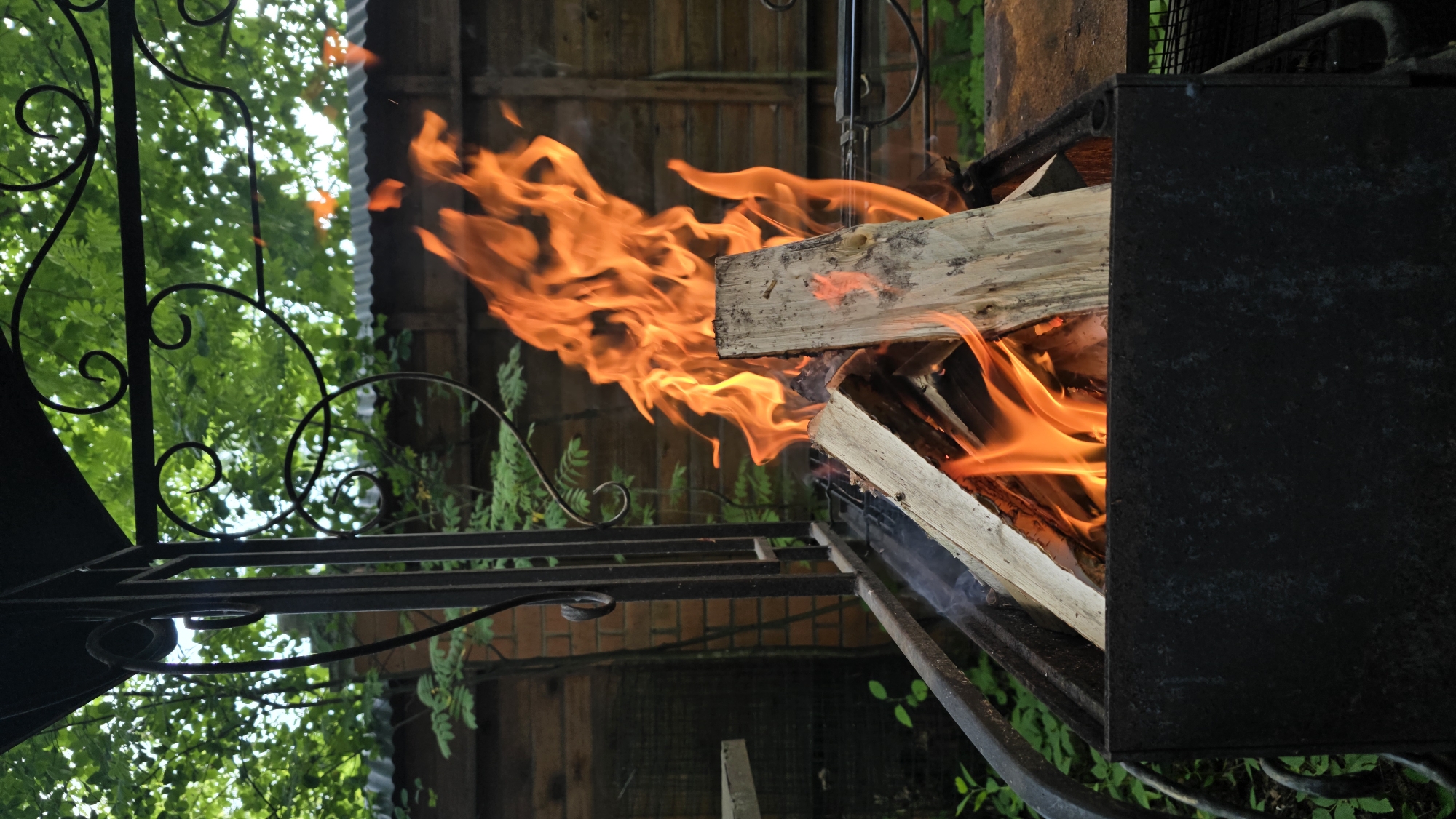 The image depicts a close-up view of a barbecue grill with burning logs in Il'inskiy, Russia. The grill, made of dark metal, contains several pieces of firewood actively ablaze with bright orange flames and wisps of smoke. The grill is situated outdoors, surrounded by lush green foliage. In the background, a wooden fence and a brick structure are visible, partially obscured by trees with dense green leaves. The overall scene suggests a warm, outdoor gathering or preparation for a meal.