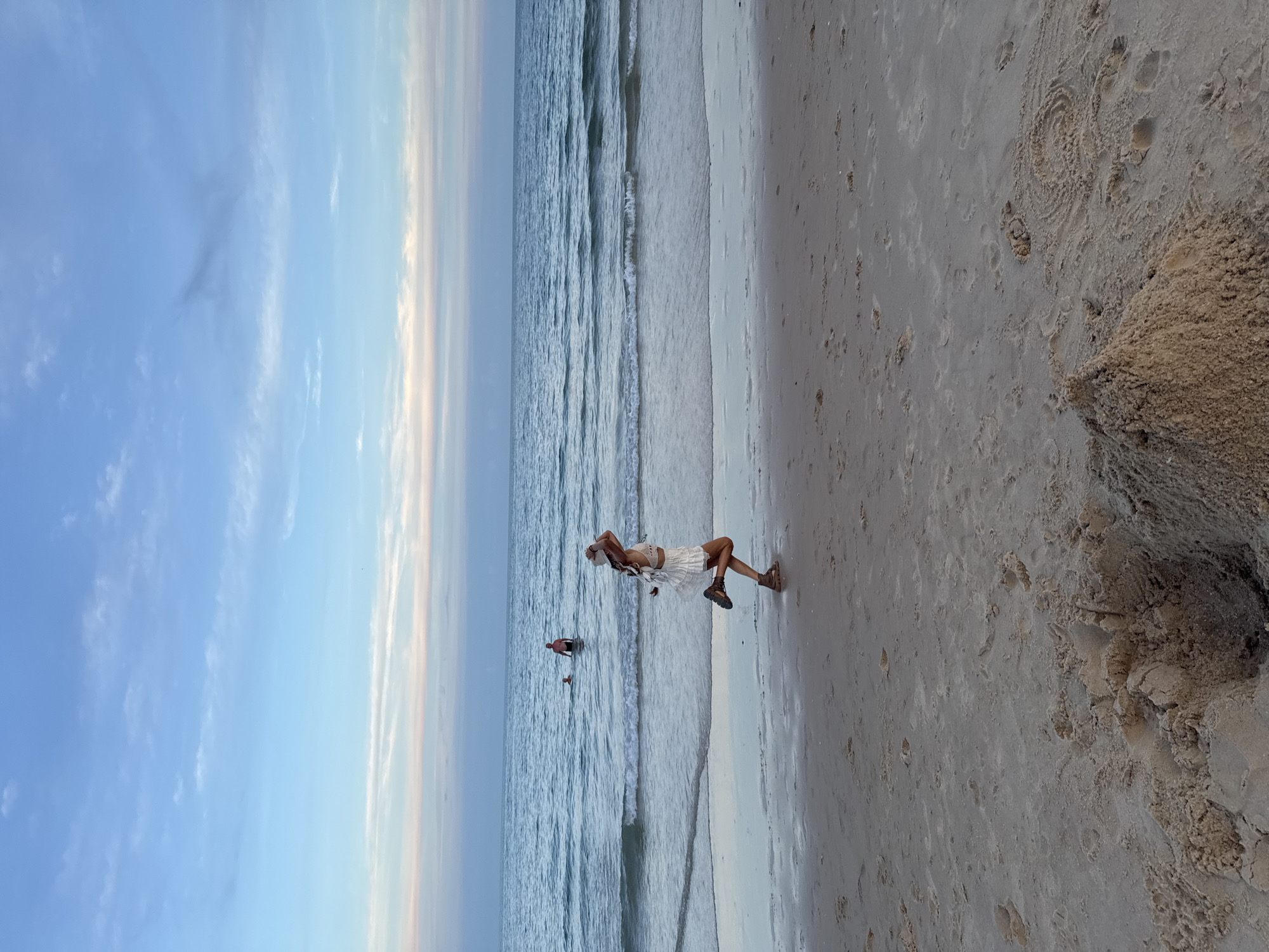 A woman in a white outfit and a hat is running on a sandy beach. She is wearing brown shoes. The ocean is behind her with gentle waves. Two men are in the water. The sky is blue with light clouds. The sand in the foreground shows footprints and a small sandcastle. The scene is likely at a beach in Phu Thuy Ward, Vietnam.