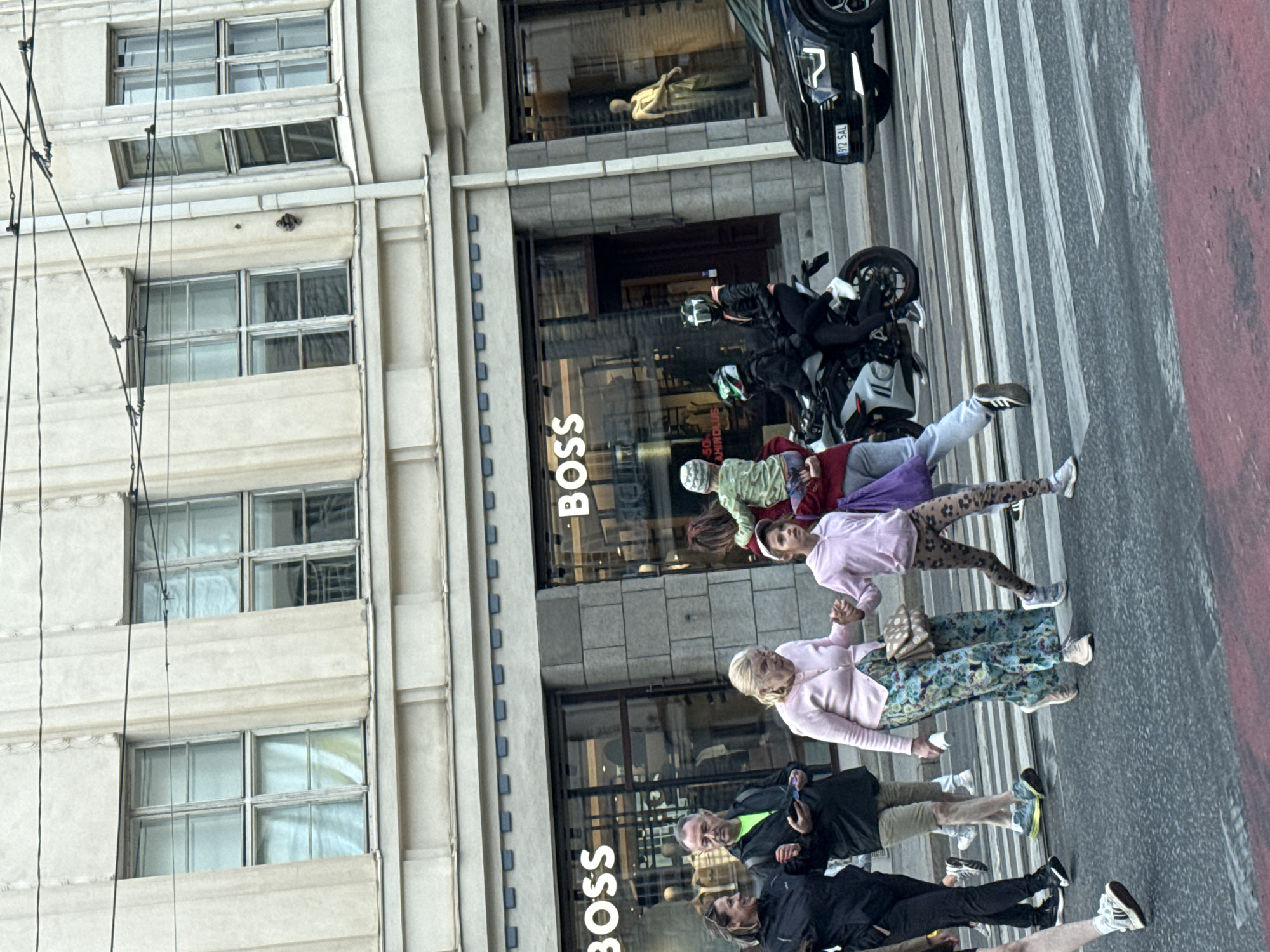In Tallinn, Estonia, a group of people are crossing a street on a zebra crossing. A woman in a black jacket walks with a man in a green shirt, holding his arm. Behind them, an older woman in a pink cardigan and a patterned skirt walks hand-in-hand with a young girl in a pink hoodie and leopard-print leggings. Another young girl, dressed in a green top and holding a baby, is being carried by a person in a red jacket, who is also walking with the group. A motorcycle with two riders wearing helmets is stopped on the street to their right, and a black car is parked behind the motorcycle. The background features a building with large windows and the logo "BOSS" displayed prominently. Overhead, power lines are visible against a light-colored sky.