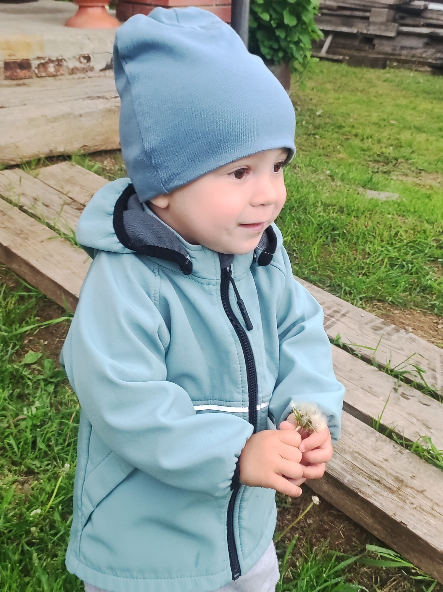 A young child is depicted outdoors, positioned slightly left of center, gazing horizontally to the right. The child wears a light periwinkle blue knitted beanie and a teal-blue softshell jacket featuring a dark grey fleece-lined collar with black trim and a black central zipper with a black toggle. The child's small hands are clasped together at mid-chest height, holding a white, spherical dandelion seed head (puffball). Light grey pants are visible at the lower edge of the jacket.
The child stands on a surface comprising green grass interspersed with patches of dark earth. To the child's right and extending into the mid-ground, two weathered, light brown wooden planks run parallel, forming a linear ground-level element. In the upper left background, a fragment of a reddish-orange ceramic pot is visible next to a light grey concrete or stone structure displaying areas of brown and orange discoloration, indicative of weathering or staining. The background on the right features green foliage and indistinct dark, stacked elements, possibly lumber or building materials. The setting is consistent with an outdoor area in Фоки, Russia.